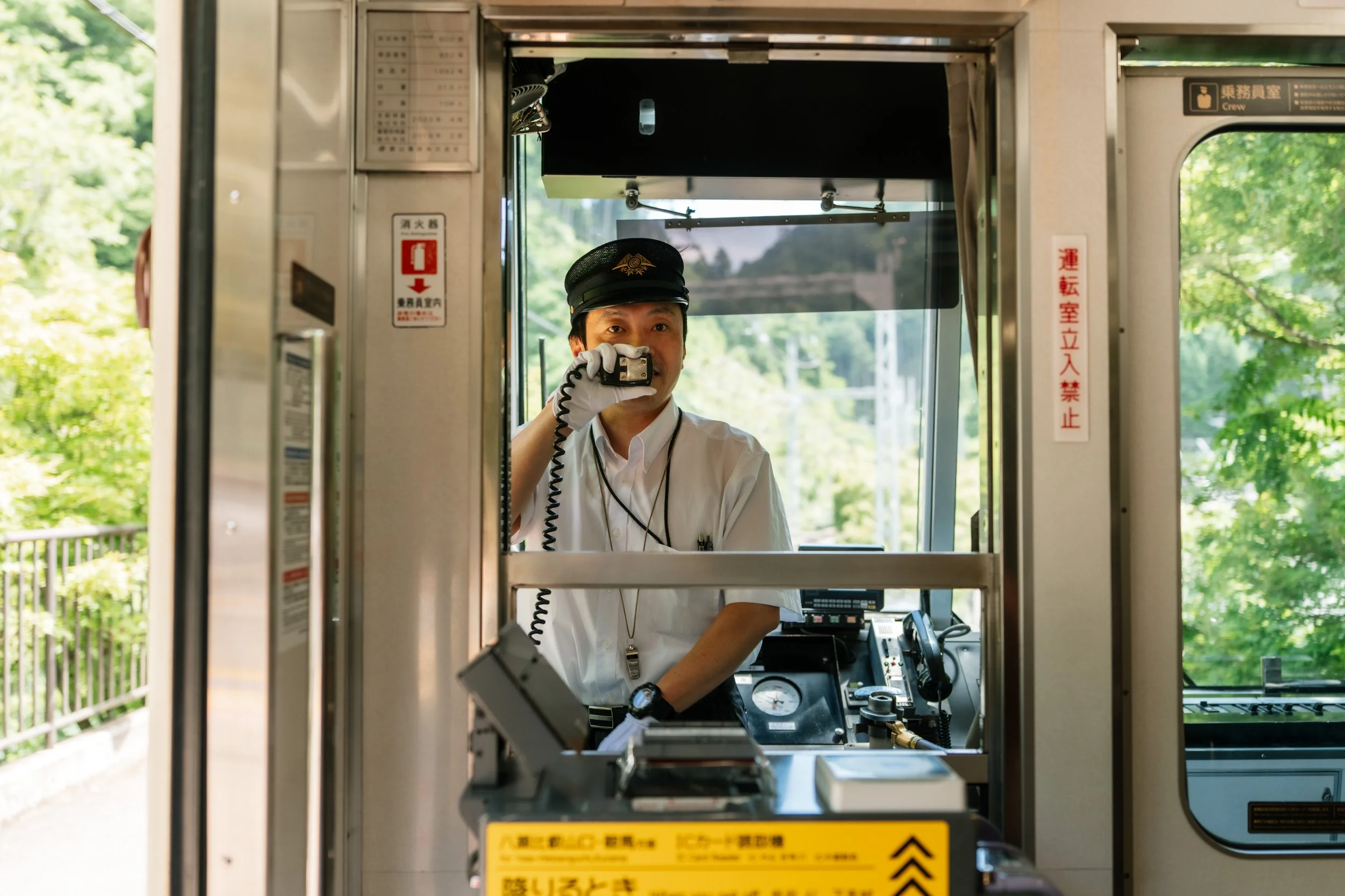A train conductor in uniform holds a radio inside a train control booth, with greenery visible outside.
