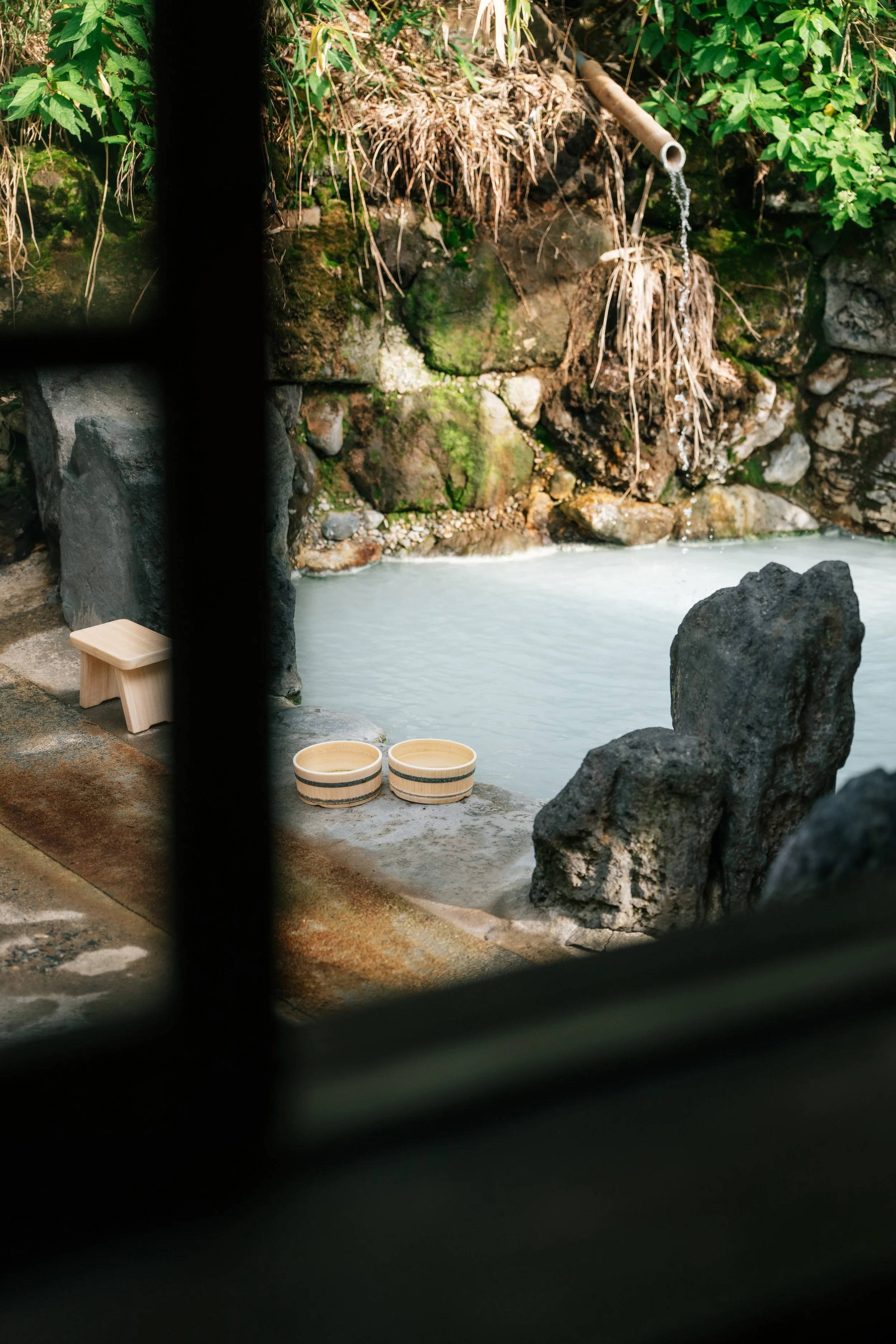 Outdoor hot spring bath (onsen) with steaming water, surrounded by rocks and lush green plants, viewed through a window with dark bars.
