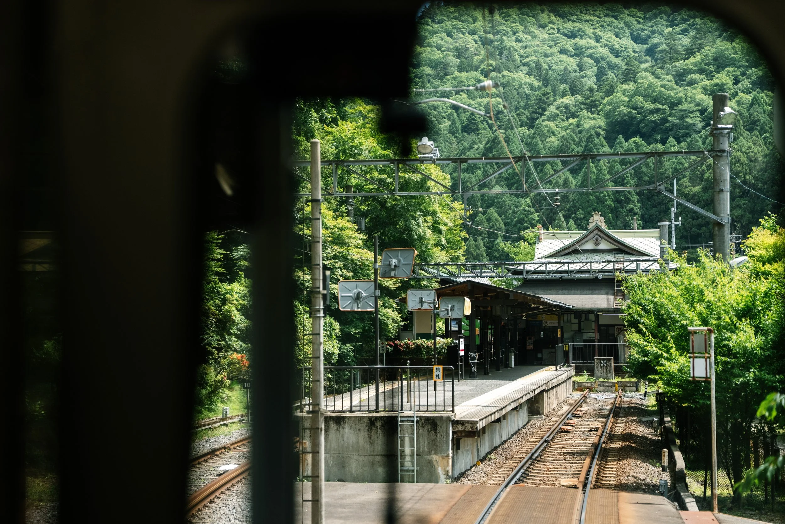 View of train tracks and a small train station surrounded by lush green trees on a hillside.