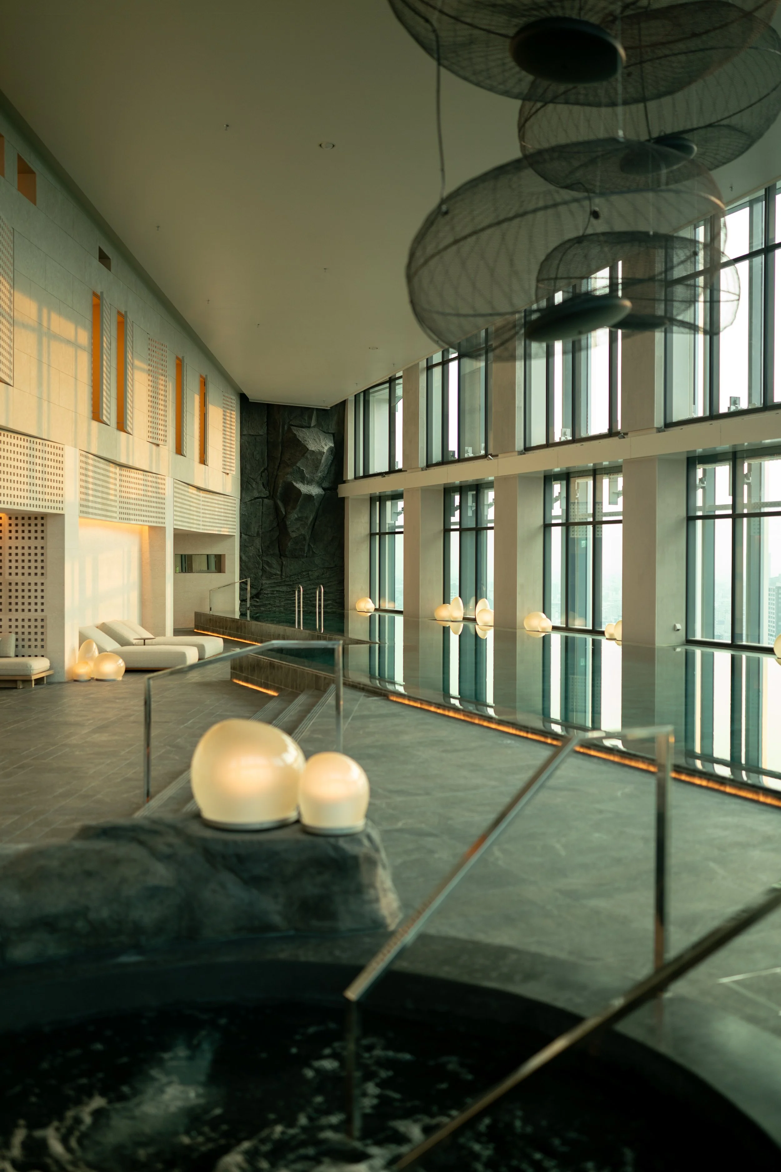 Interior view of a modern, luxurious indoor swimming pool area with large floor-to-ceiling windows, lounge chairs, spherical lamps, and artistic ceiling fixtures at JW Marriott Tokyo hotel, editorial hospitality photography, Japan.
