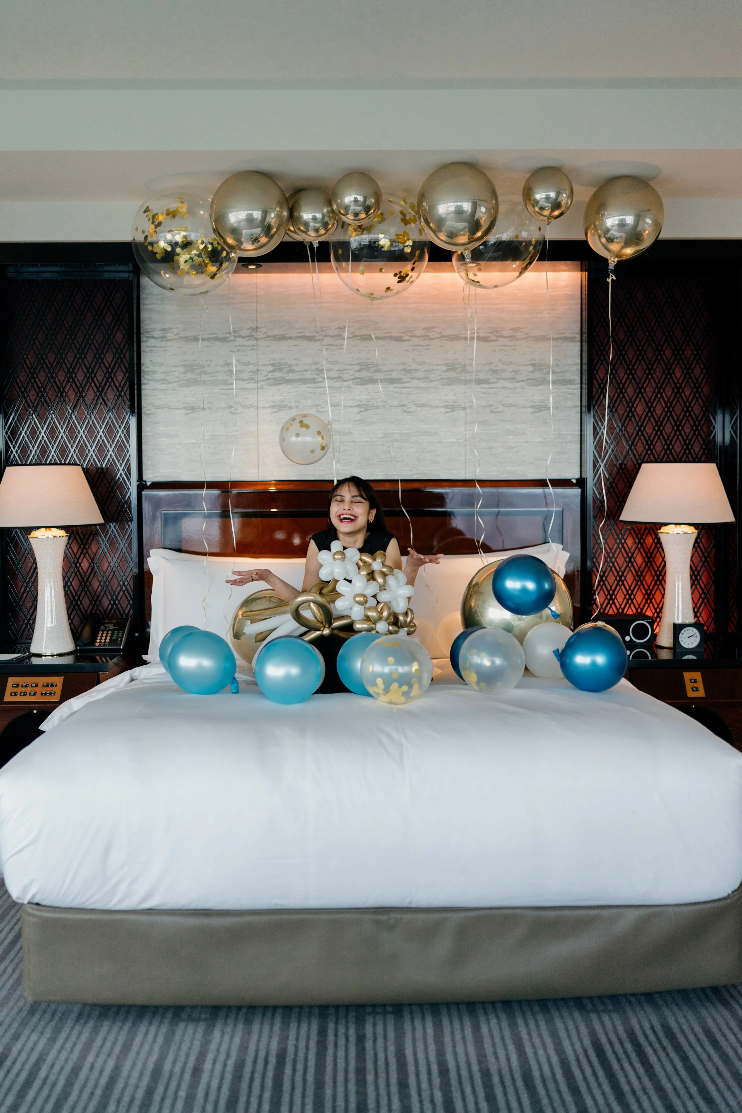 A woman sitting on a bed surrounded by gold, silver, and blue balloons, with more balloons hanging from the ceiling, in a hotel room with lamps and a landscape painting of water in the background.
