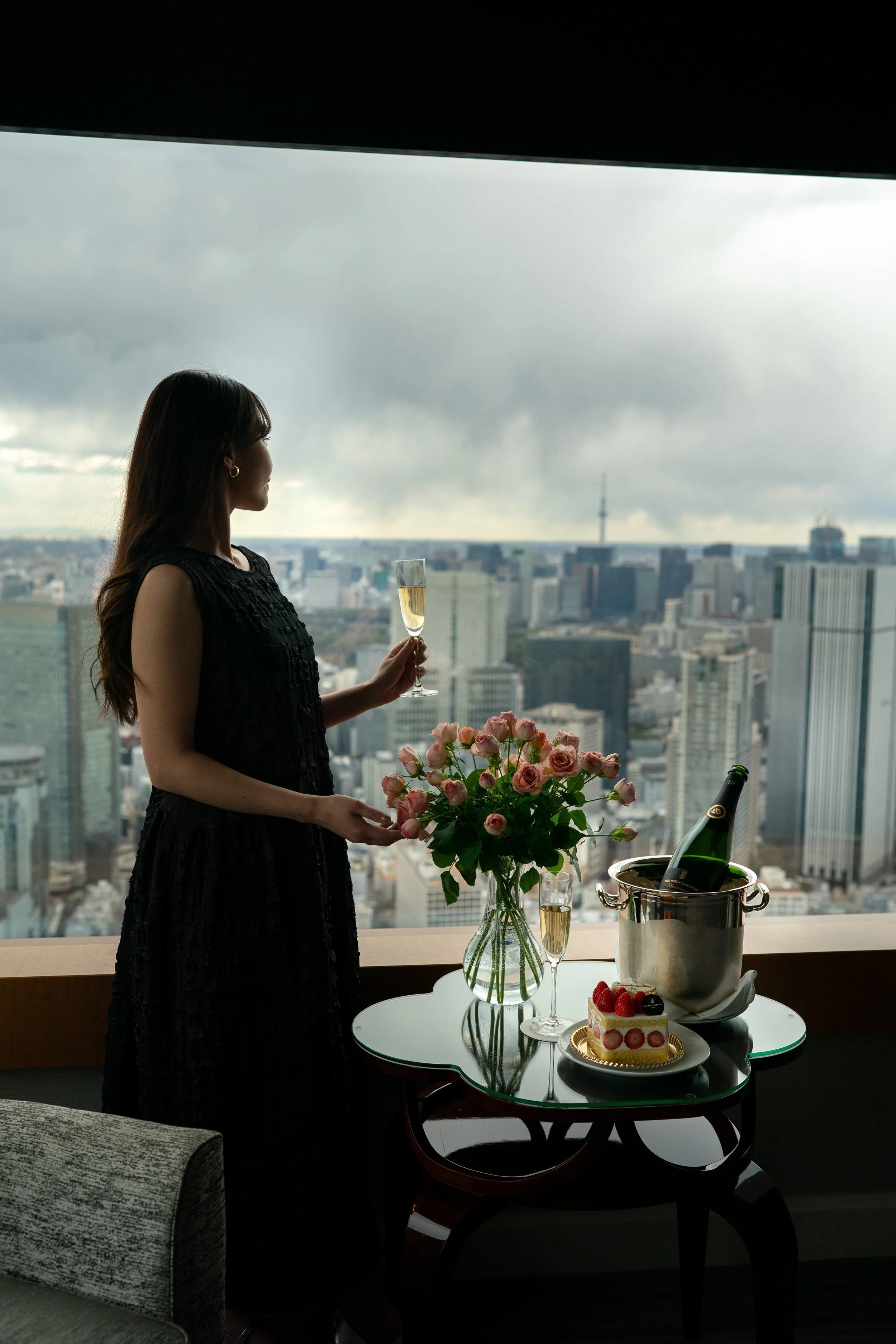 A guest with long brown hair in a black dress holding a glass of champagne with a city skyline view, a table with flowers, a cake, and a champagne bucket at The Ritz-Carlton, Tokyo. Editorial hotel photography, Japan.