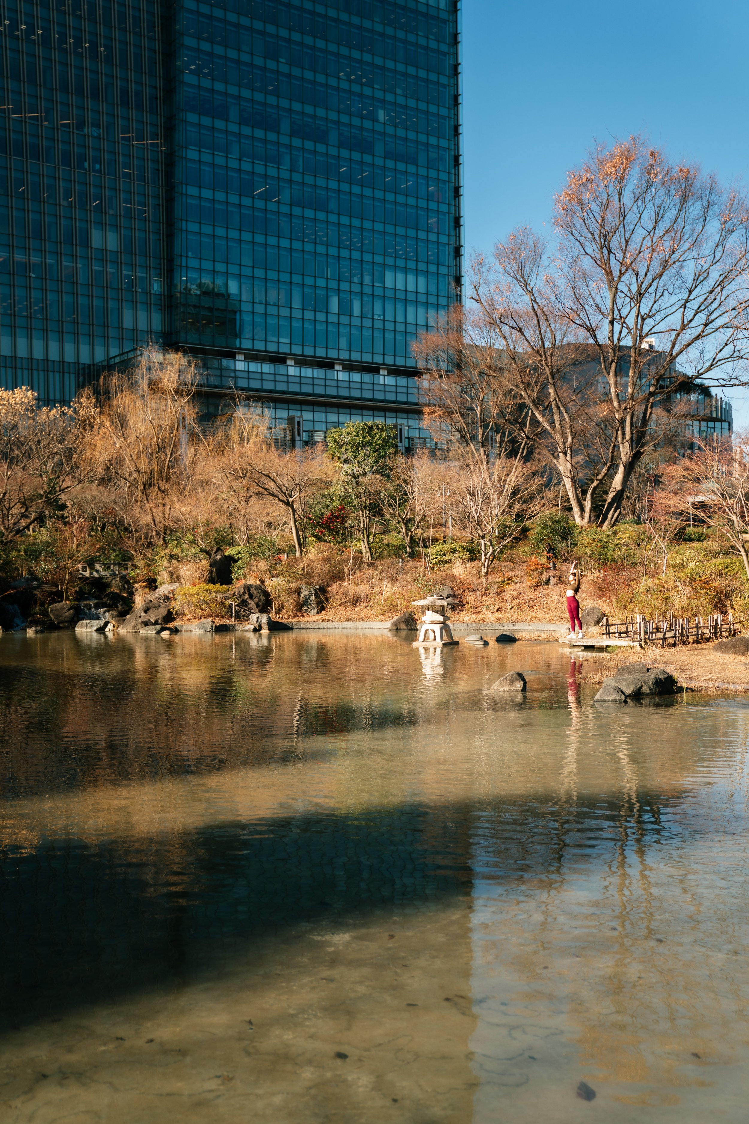 A modern glass skyscraper where The Ritz-Carlton, Tokyo is located. A pond with a person standing on a small path beside it. Editorial hospitality photography showcasing guest wellness experience.