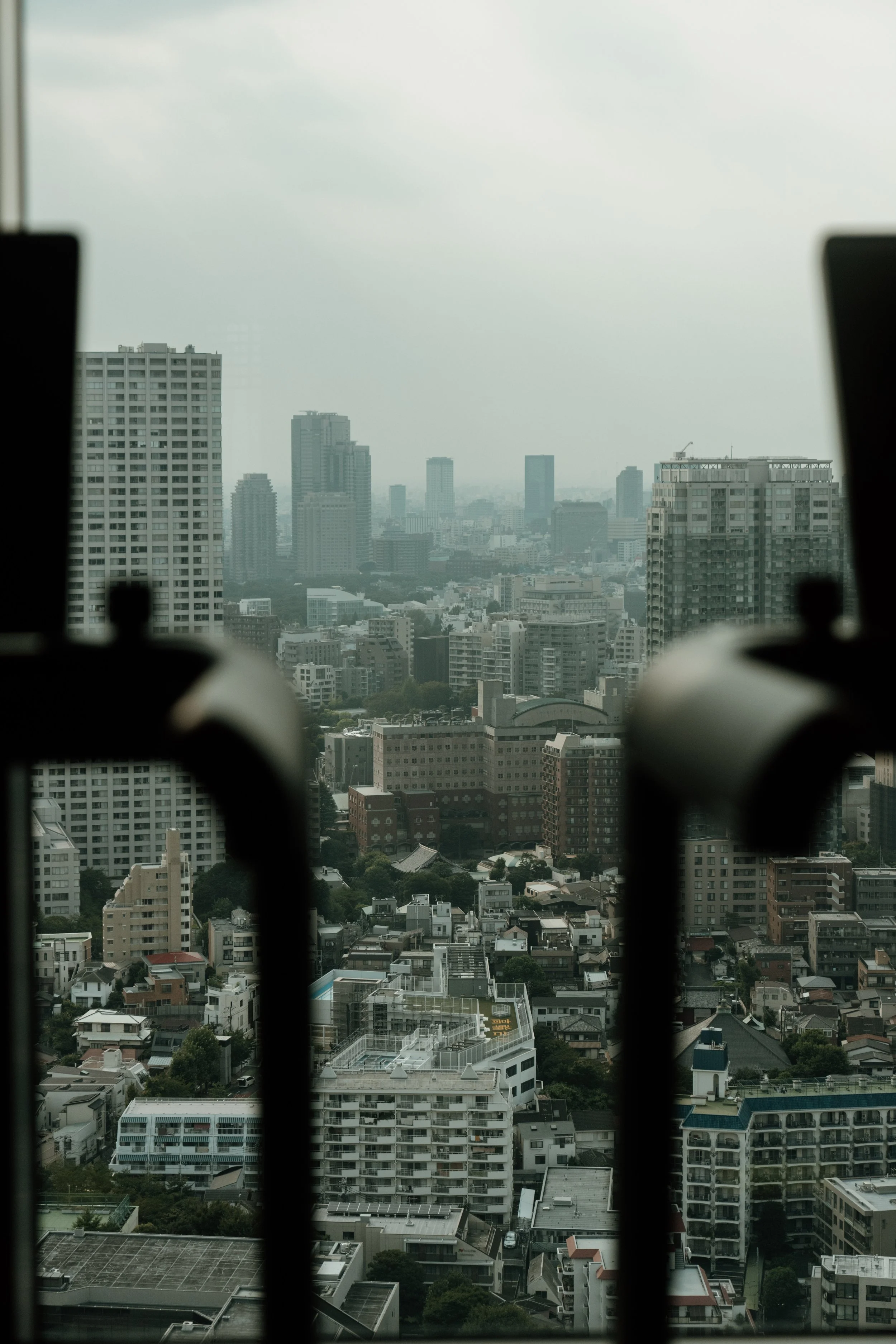 Cityscape view of high-rise buildings and residential apartments through window with black railings in the foreground under a cloudy sky at JW Marriott Tokyo hotel, editorial hospitality photography, Japan.