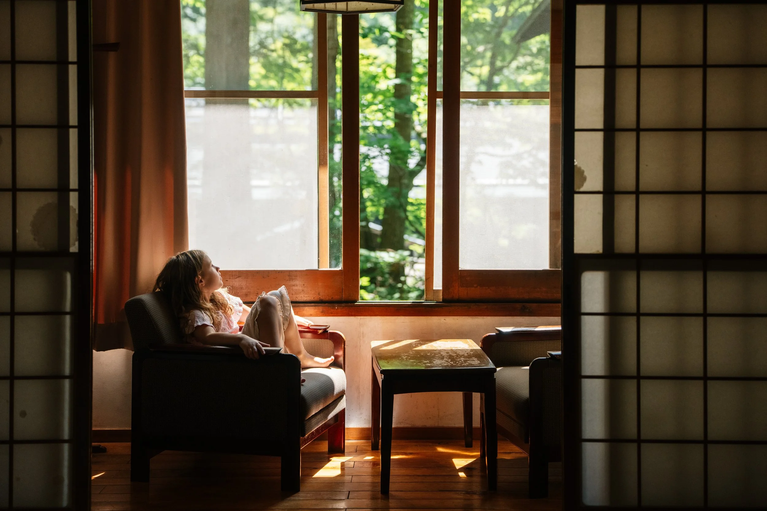 A young girl relaxing on a chair in a sunlit room with large windows showing green trees outside, framed by shoji screens.
