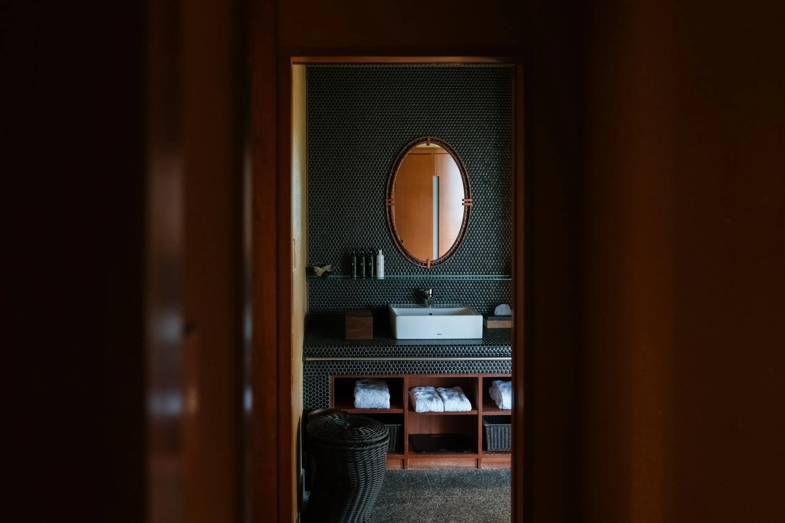 A bathroom view through a doorway showing a black tiled wall with an oval mirror, a white sink, the bottom of a towel rack, and shelves with towels.