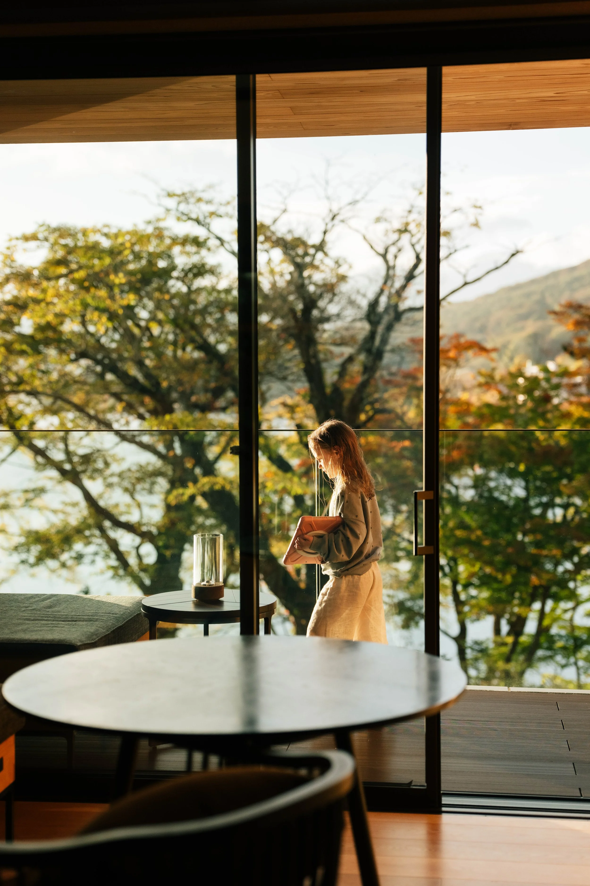 A woman with long hair standing inside a modern building near a large glass window with a railing outside showing trees with fall foliage. There is a round table with a glass vase on it, and in the foreground, a blurred round table and chairs.