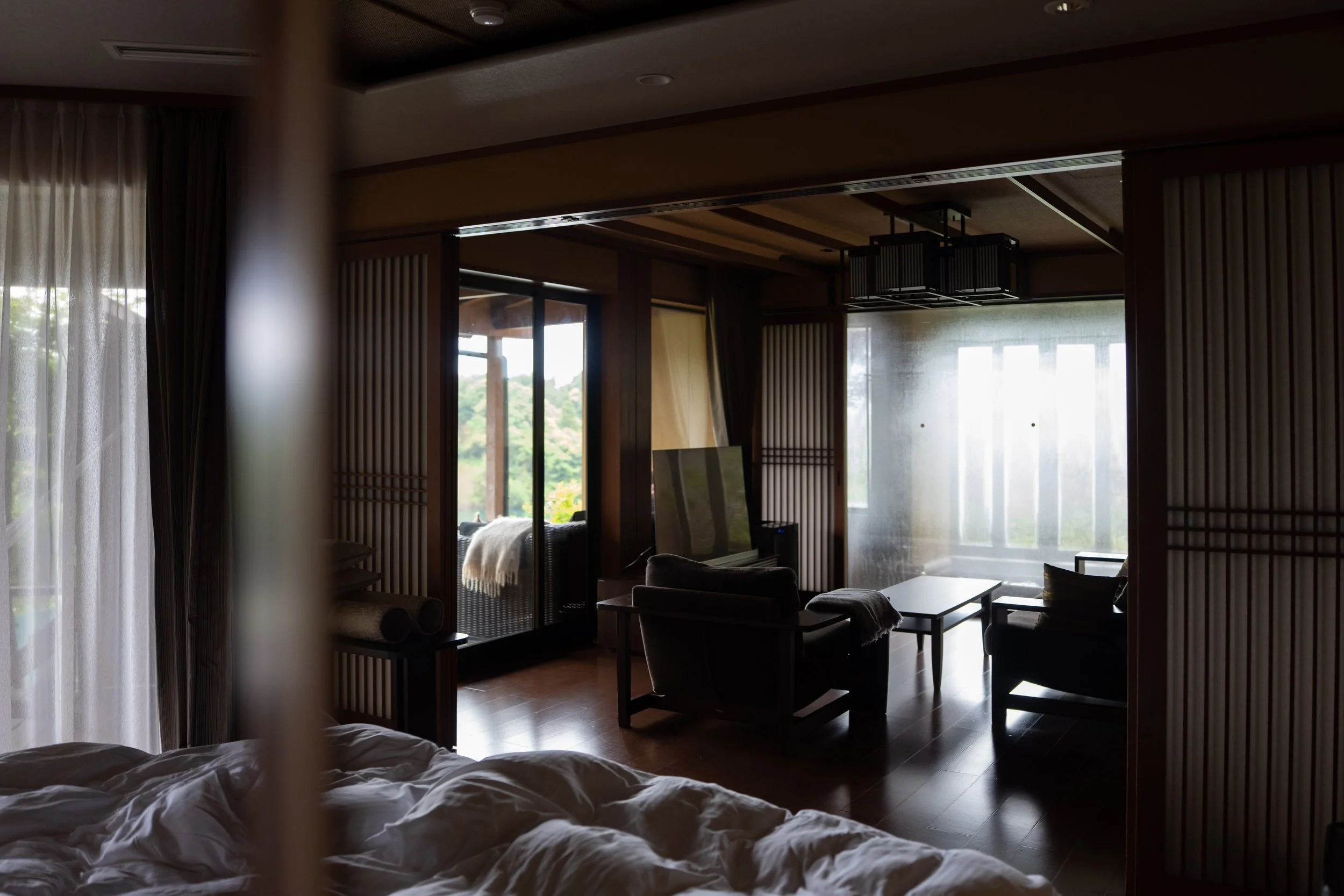 A dimly lit room with a bed in the foreground, white bedding, and large windows with curtains letting in natural light.