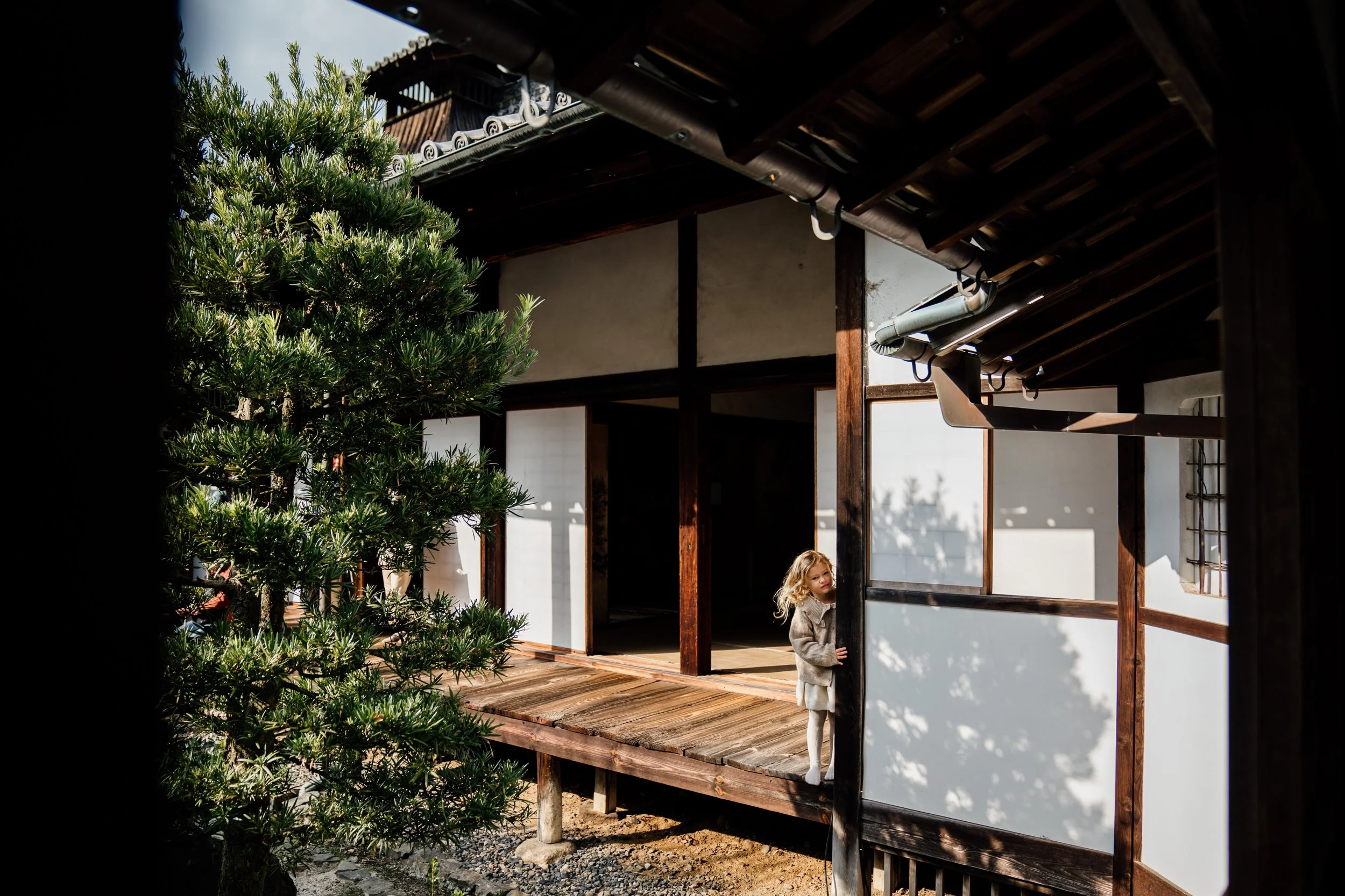 A young girl in a gray coat standing on a wooden porch, partially hiding behind a wall, with a traditional Japanese house and lush green trees in the background.