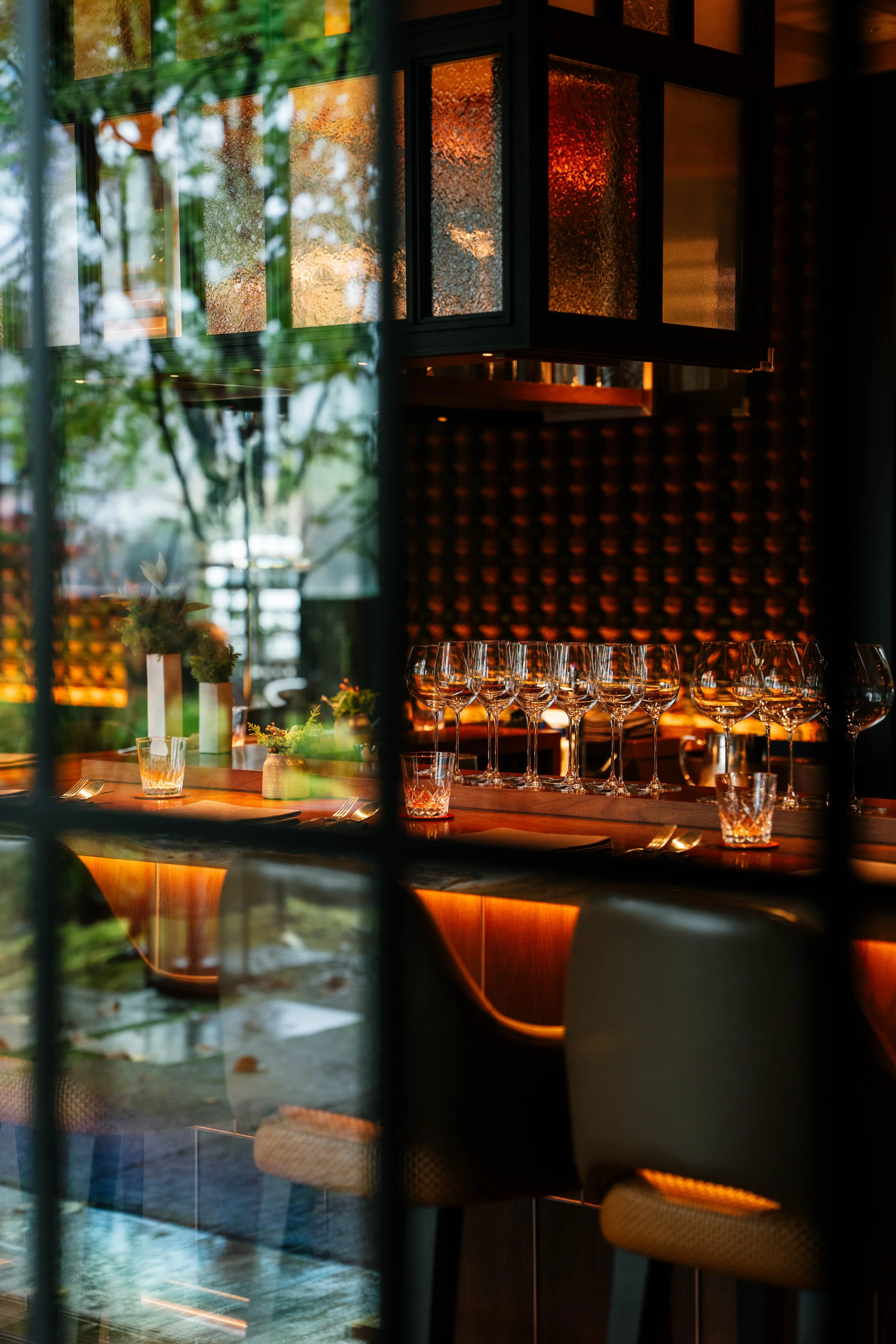 Interior of a restaurant or bar, with a row of empty wine glasses on the bar counter, illuminated by warm lighting, seen through a window with reflections of trees outside.