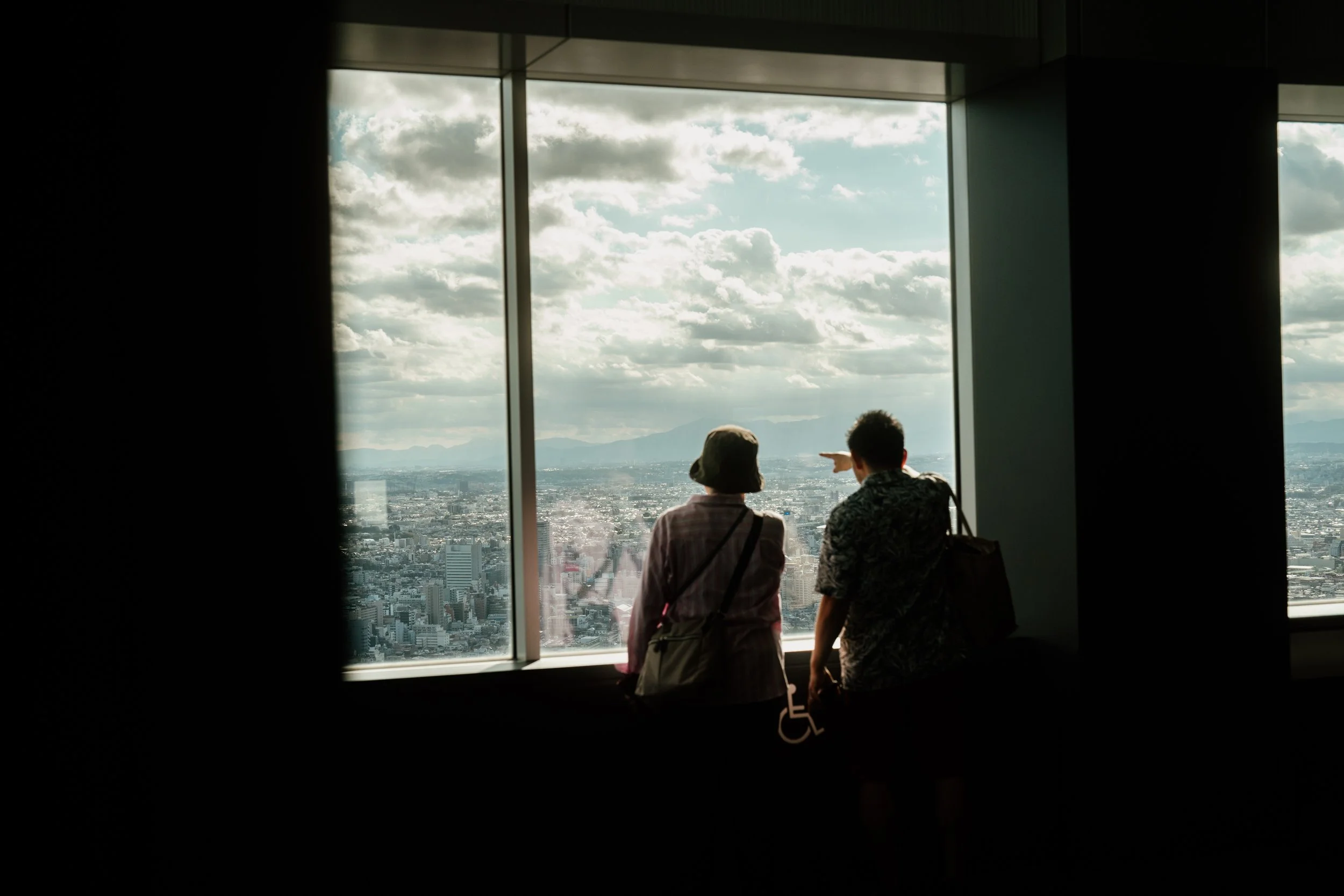 Two people, one with a hat, looking out a large window of a high-rise building at a cityscape and cloudy sky. One person is pointing toward the view.
