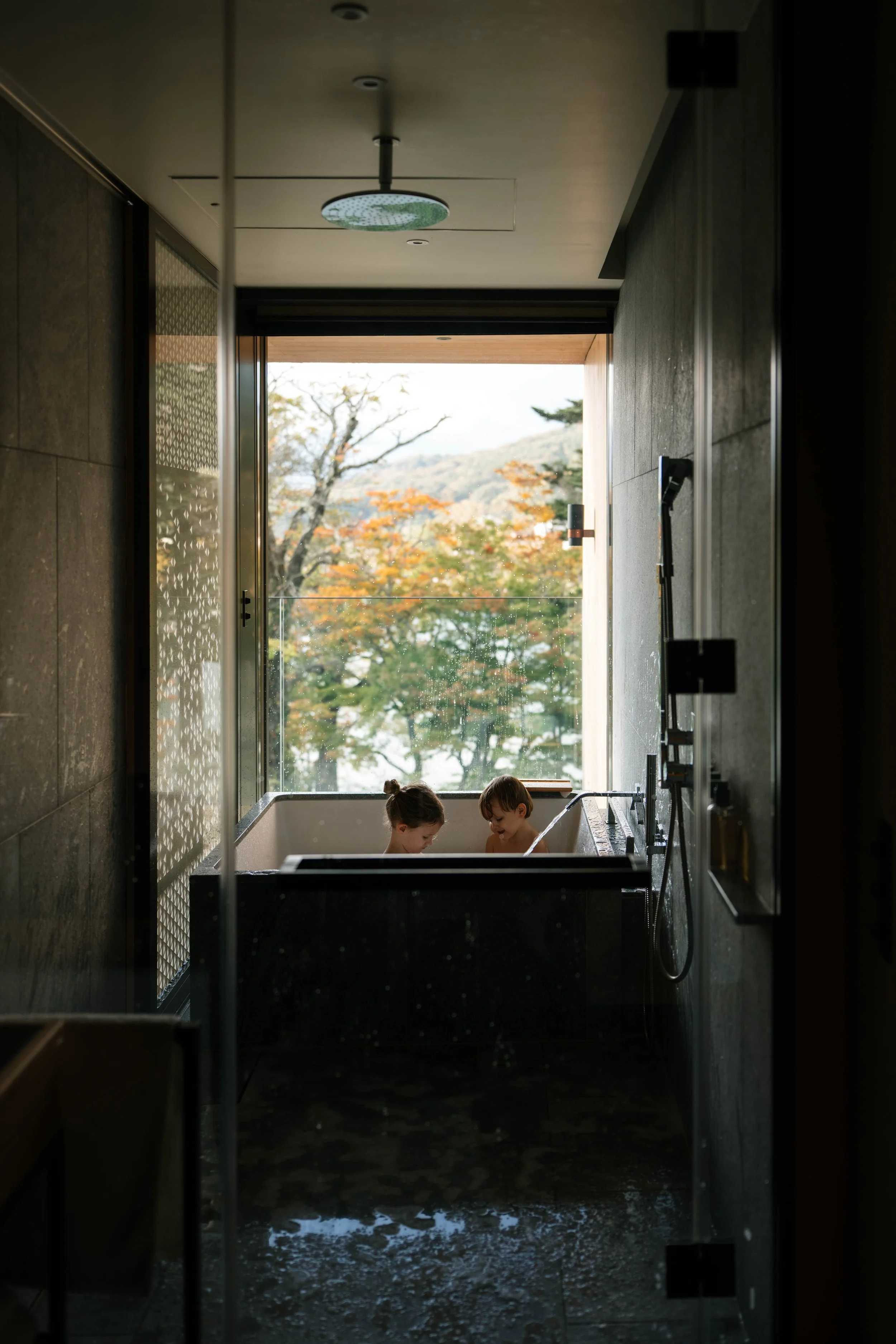 Two children taking a bath in a black bathtub near a large window with an autumn landscape outside, featuring trees with orange and yellow leaves.