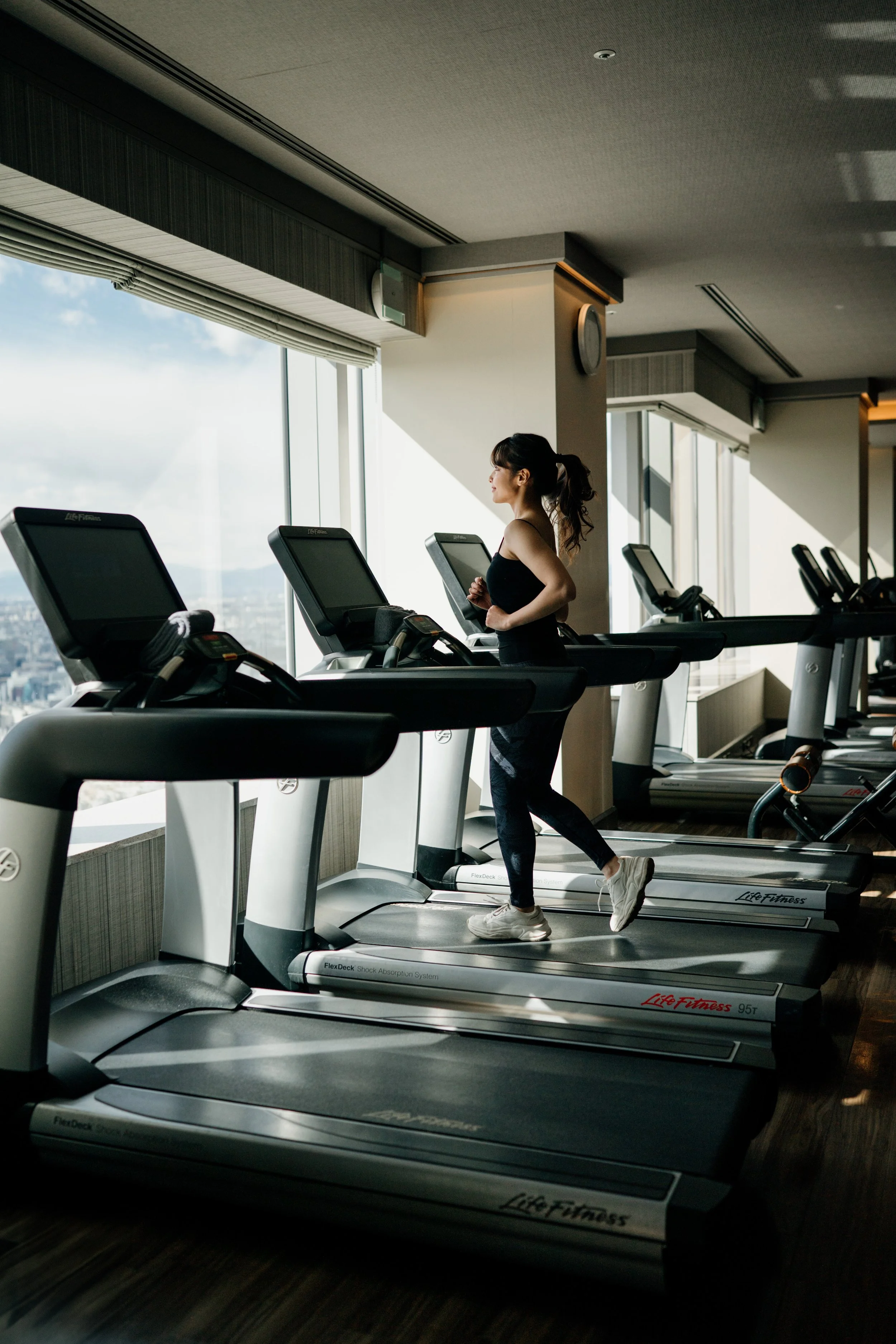A woman is running on a treadmill in a gym with large windows showing a city view outside. The Ritz-Carlton, Tokyo, editorial hospitality photography, Japan,  showcasing guest wellness experience. 