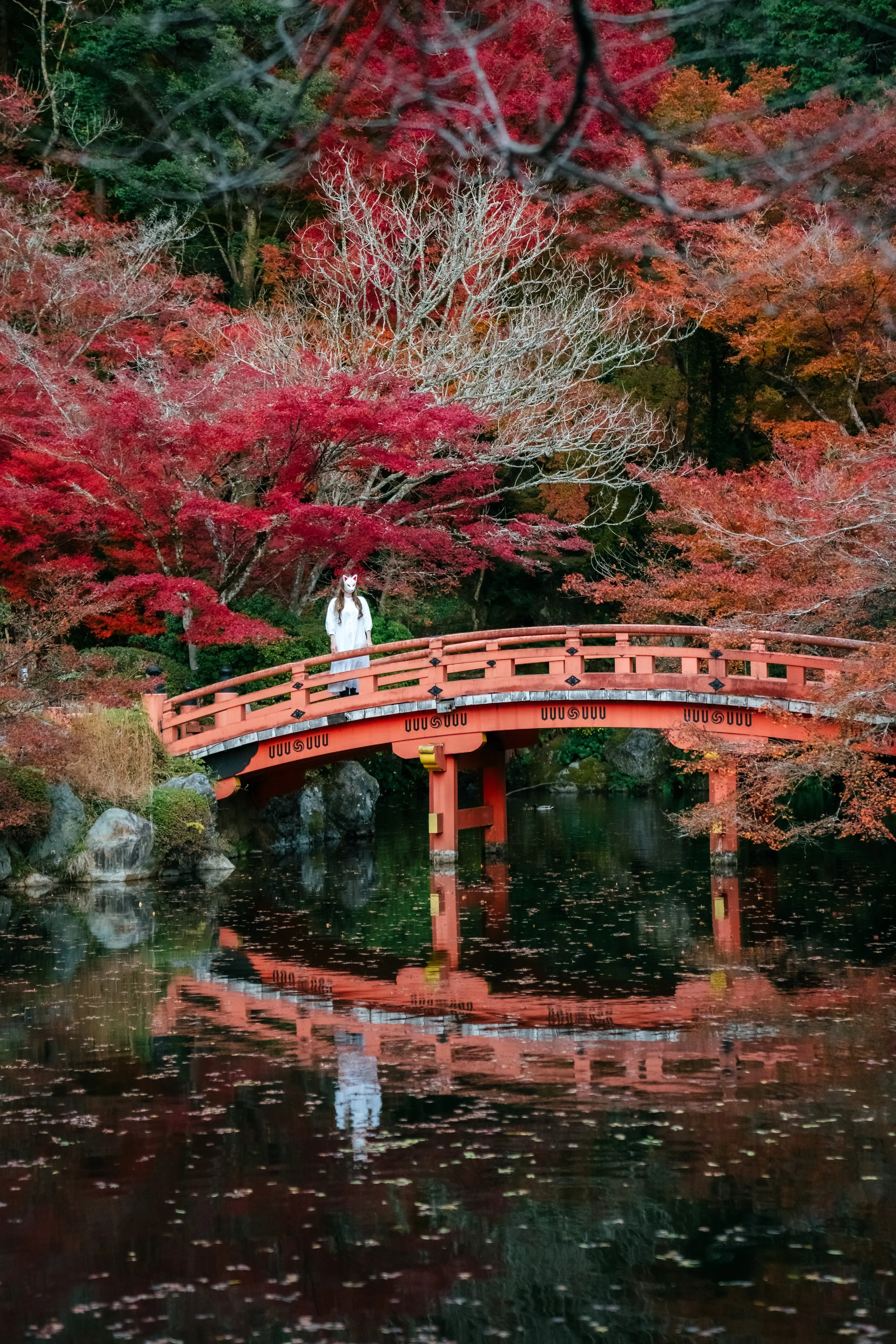 A person wearing a white mask and coat standing on a red bridge surrounded by colorful autumn trees, reflected in the water below.