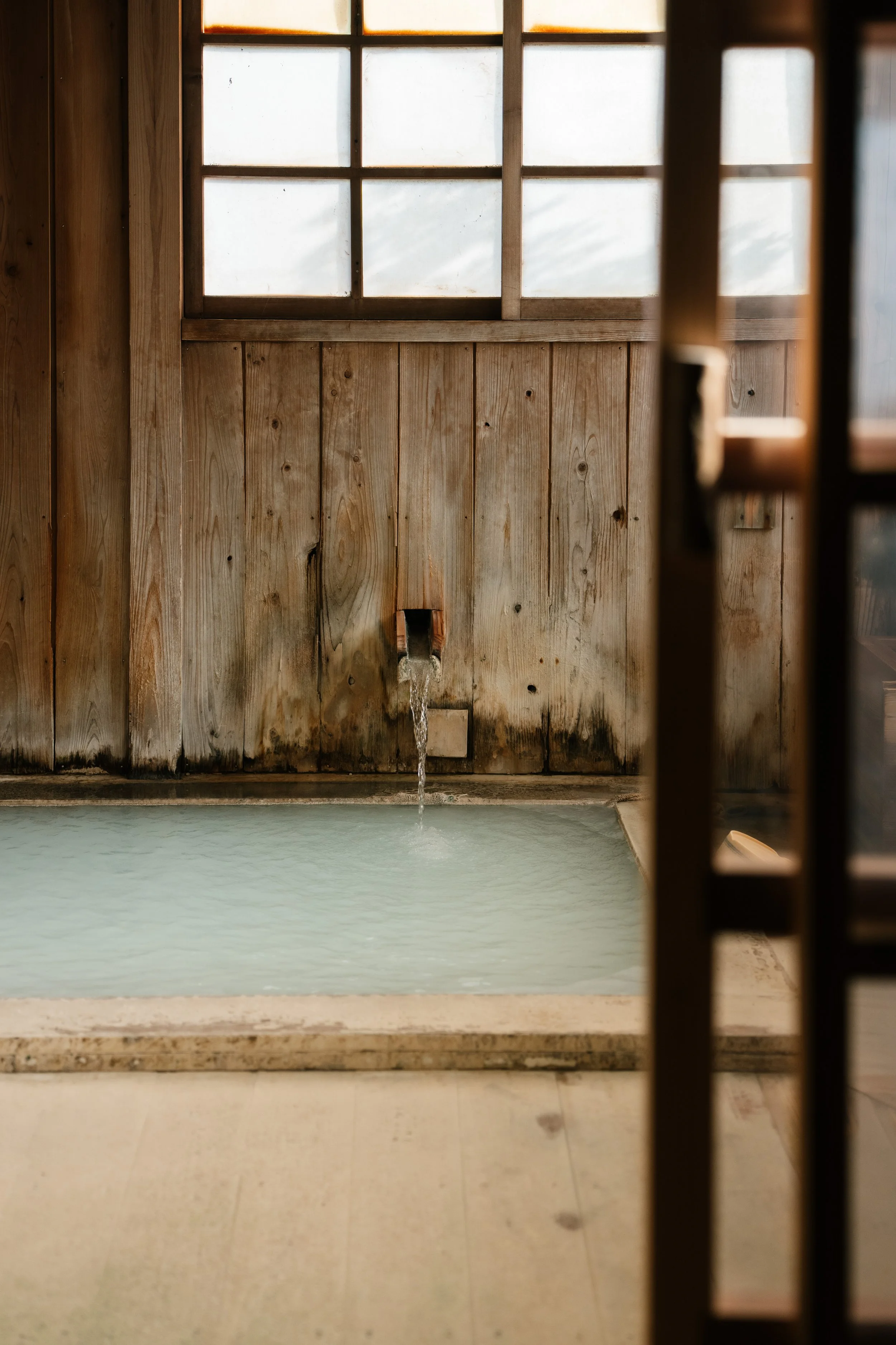 A wooden interior with a window on the upper part and a water feature below, where water flows out from a pipe into a pool, framed by dark wooden posts.