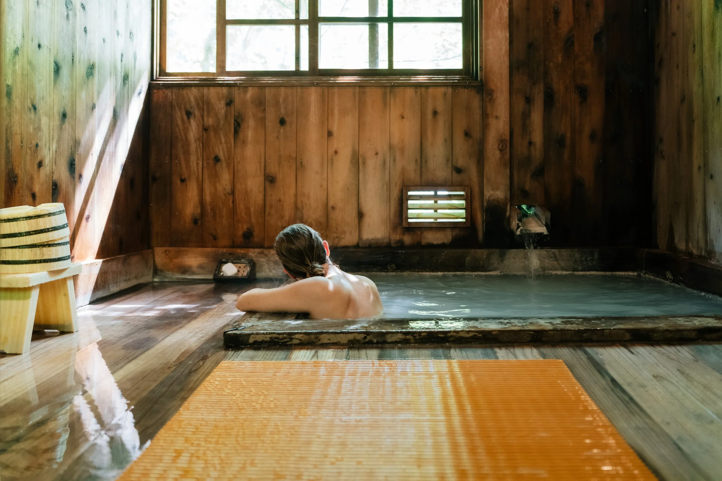 Person relaxing in a wooden indoor hot spring or bath with wooden walls and window.