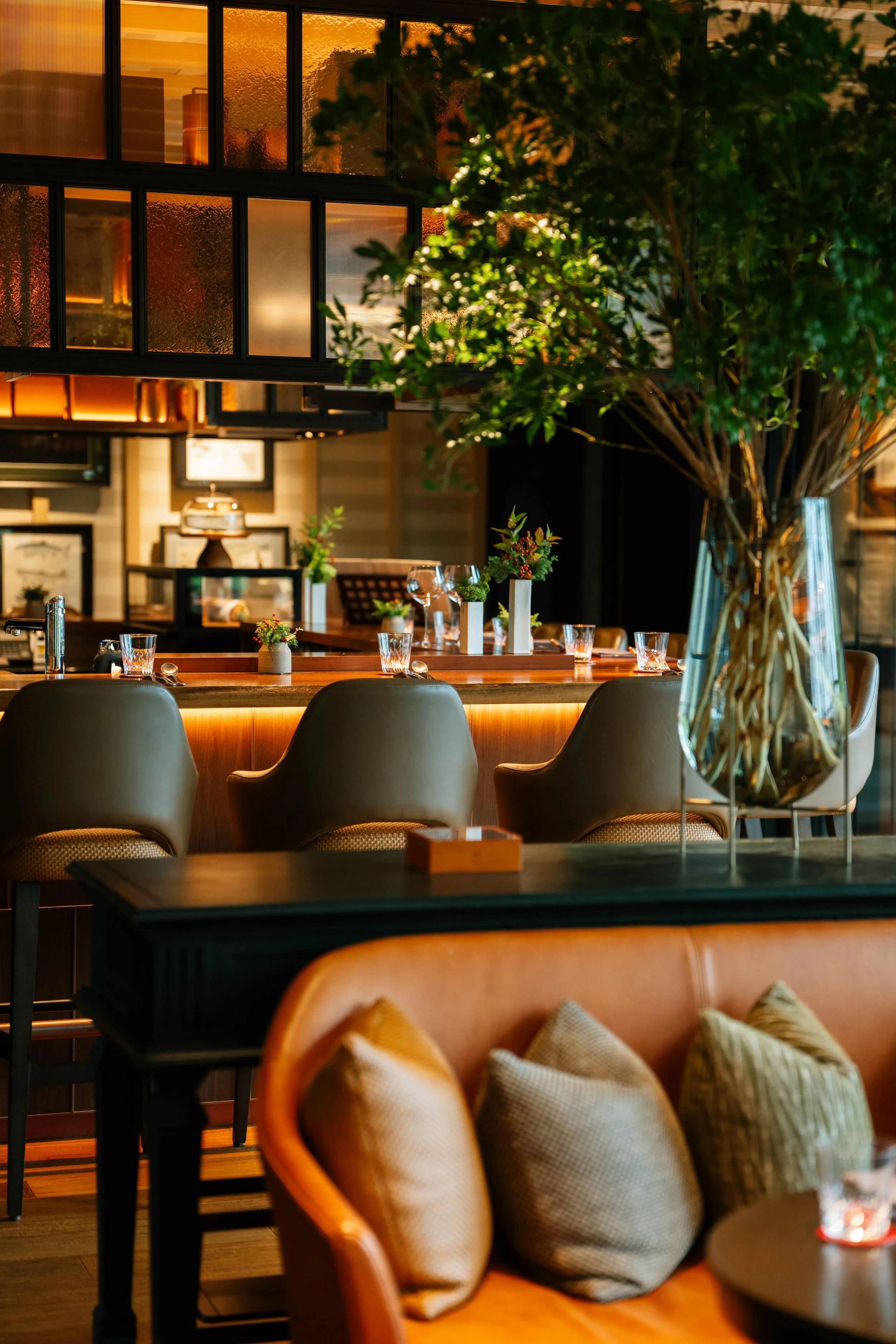 A cozy restaurant interior with a polished wooden table, beige chairs with cushions, decorative plants, and warm lighting. There are large windows with textured glass and contemporary decor.