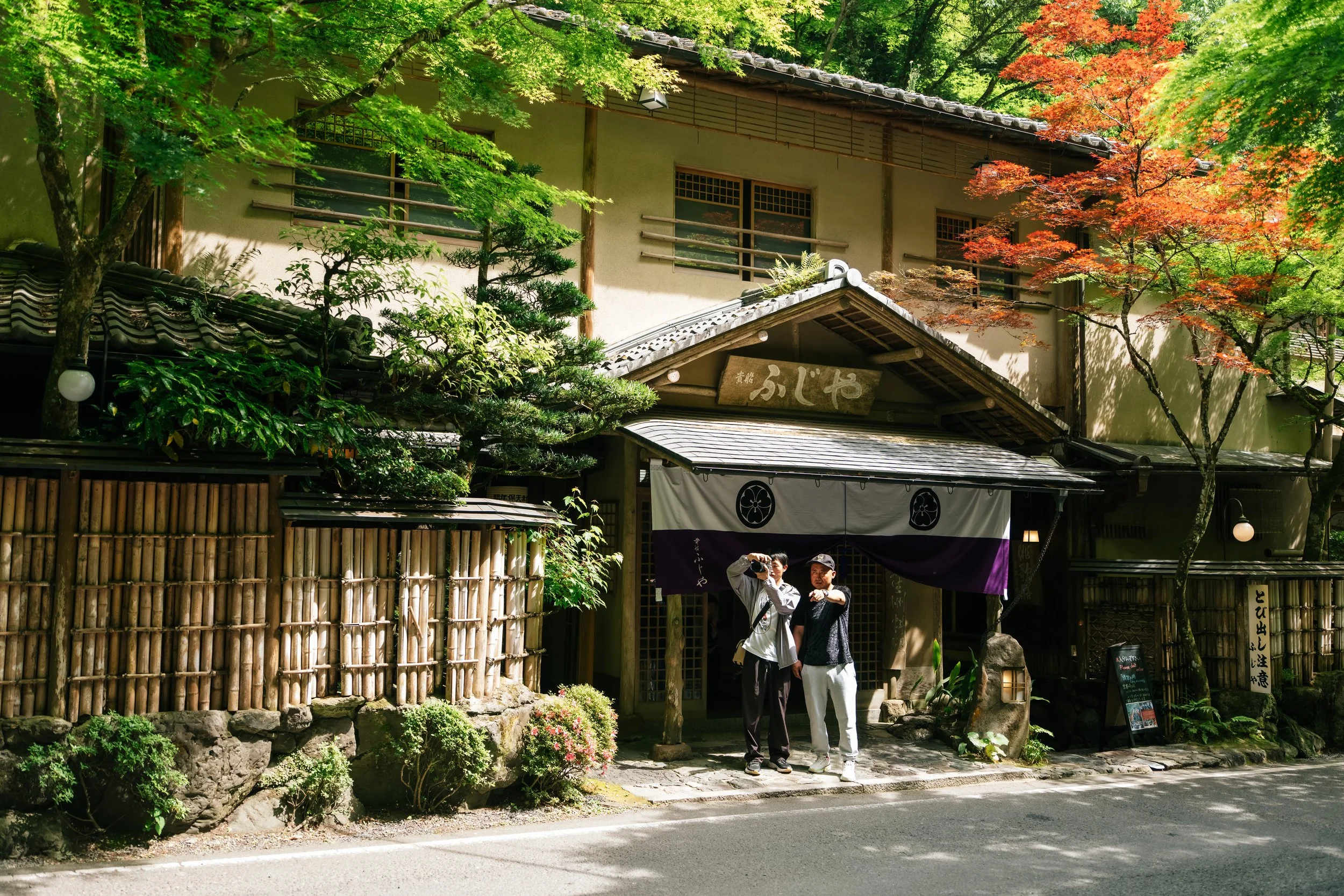 Traditional Japanese building with wooden and tiled roof, surrounded by lush green trees with some red leaves, and three people standing in front, one taking a photo.