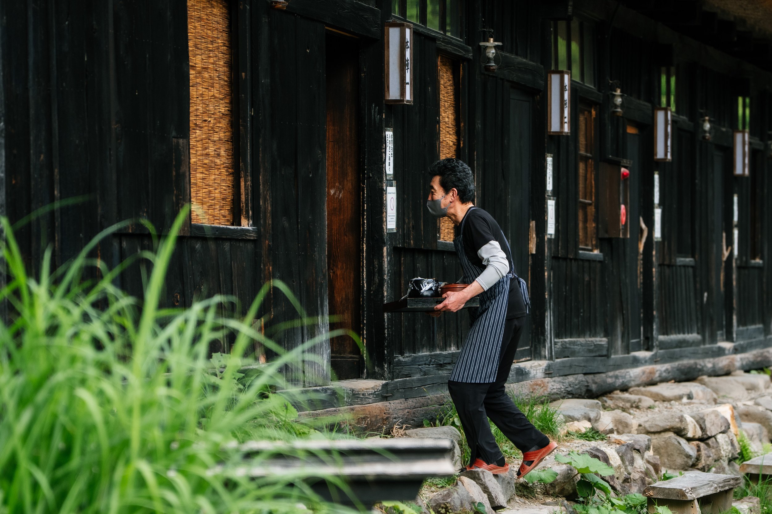 A man wearing a black face mask, black shirt, and striped apron carrying a tray of food outside a dark wooden building with hanging lanterns.