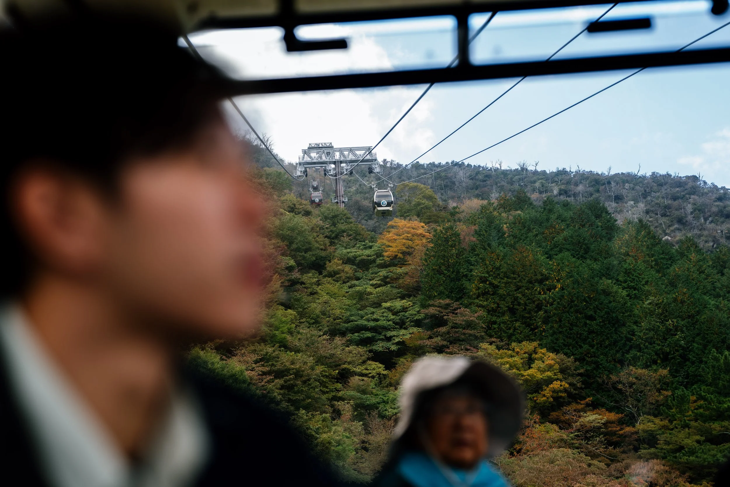 View of a cable car system over a forested area with trees showing fall colors, with people in the foreground, some partially visible and out of focus.