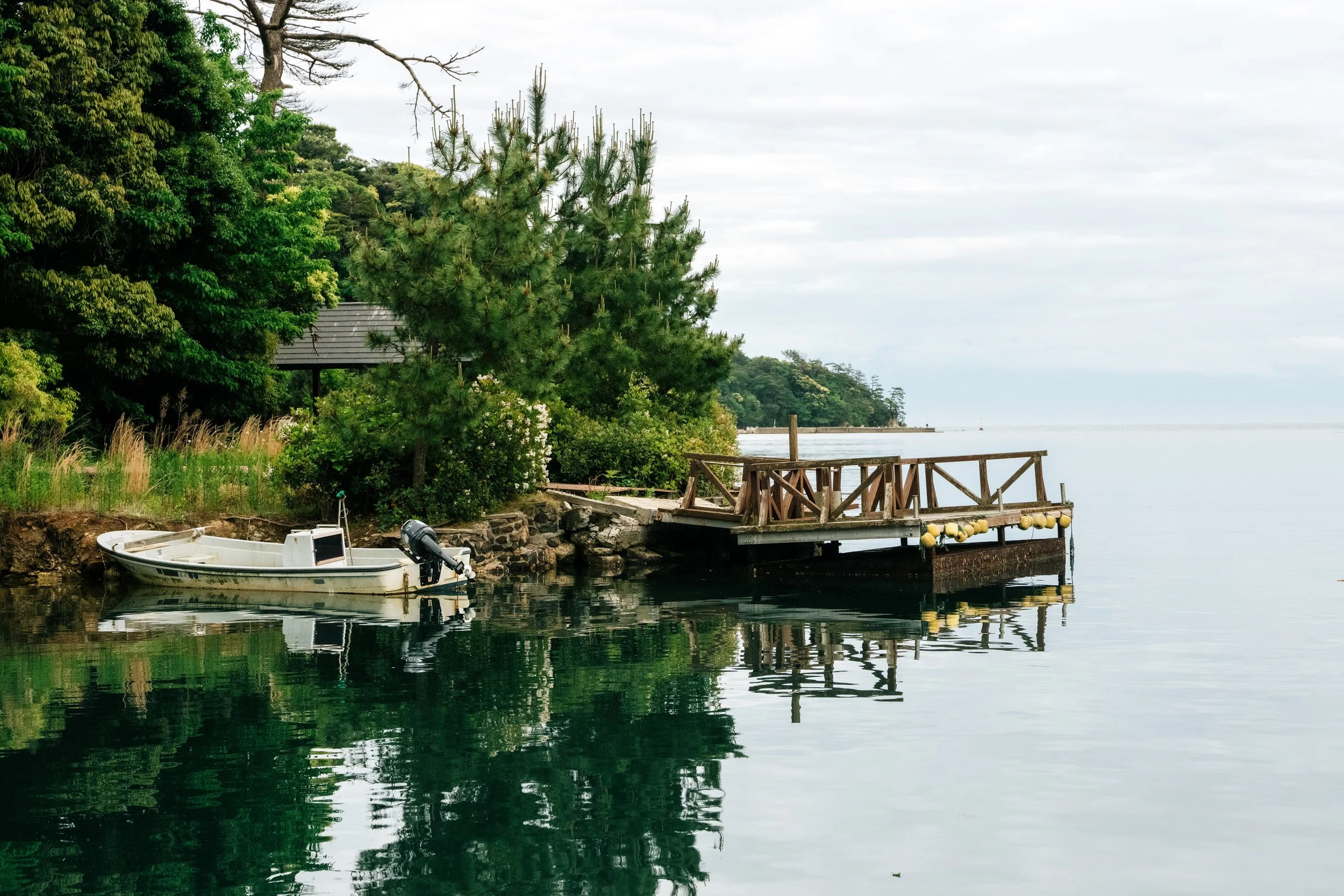 A small boat docked by a wooden pier on a calm body of water, surrounded by lush green trees and a cloudy sky.