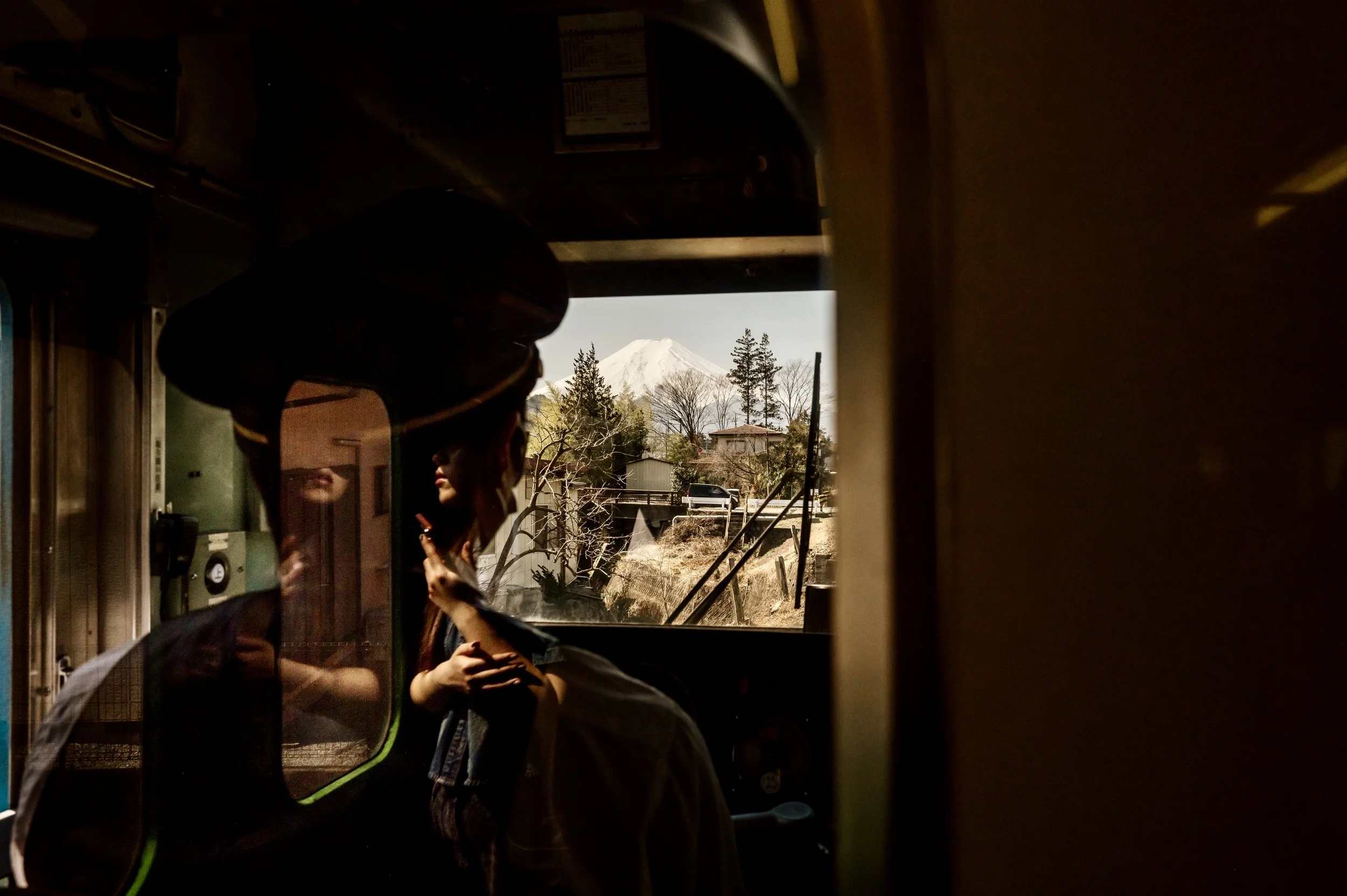 View of Mount Fuji seen from inside a vehicle. Reflection of a woman taking a photo is visible in a mirror, with trees and buildings in the background.