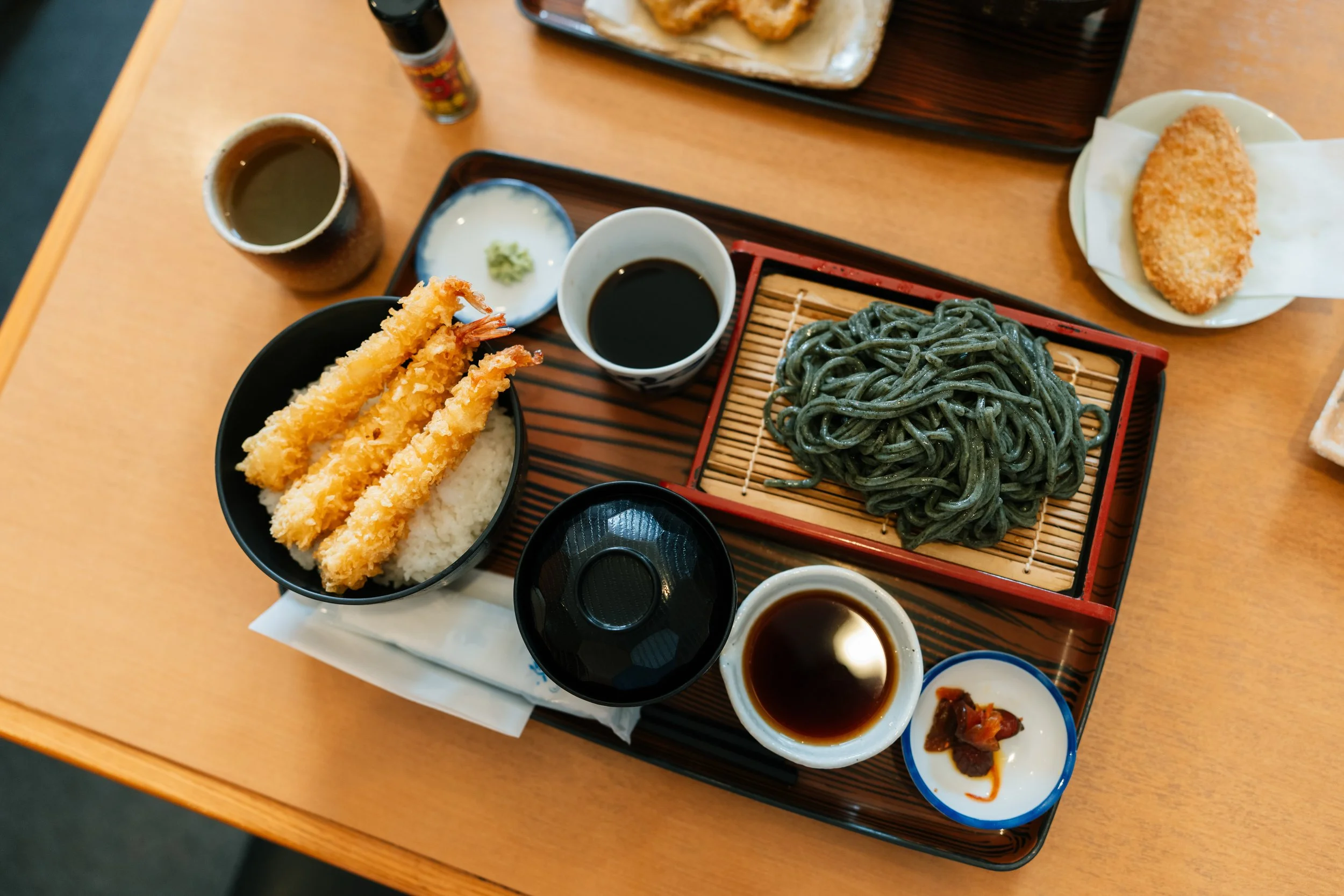A Japanese meal set on a wooden tray with tempura shrimp, cold soba noodles, dipping sauces, wasabi, pickles, and a bowl of rice.