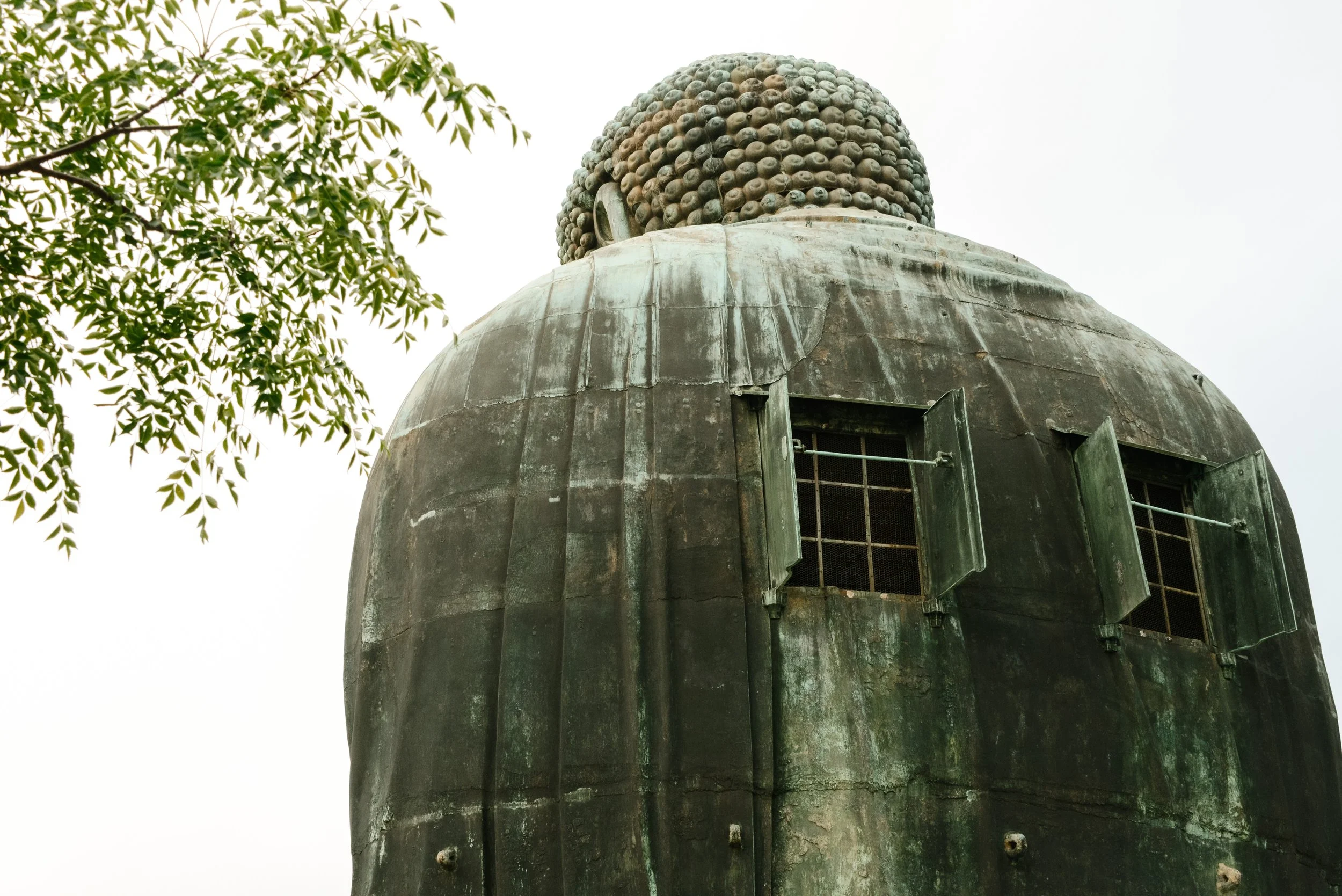 Upward view of a weathered bronze or copper statue with a rounded, beaded head and an open window-like opening on the back, surrounded by green leaves and a gray sky.
