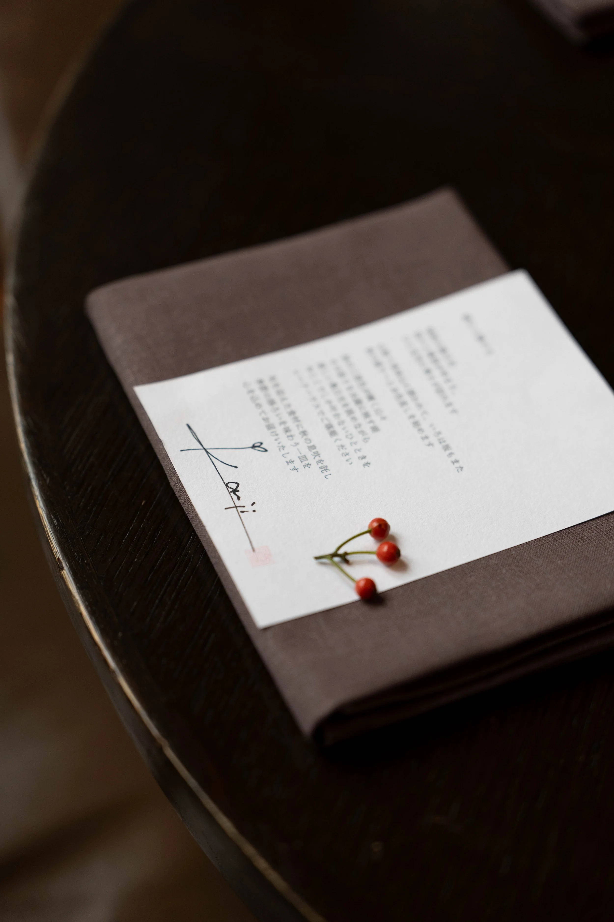 A white piece of paper with handwritten Japanese characters and a signature in black ink, placed on a brown textured mat with three small red berries nearby, on a dark wooden table.