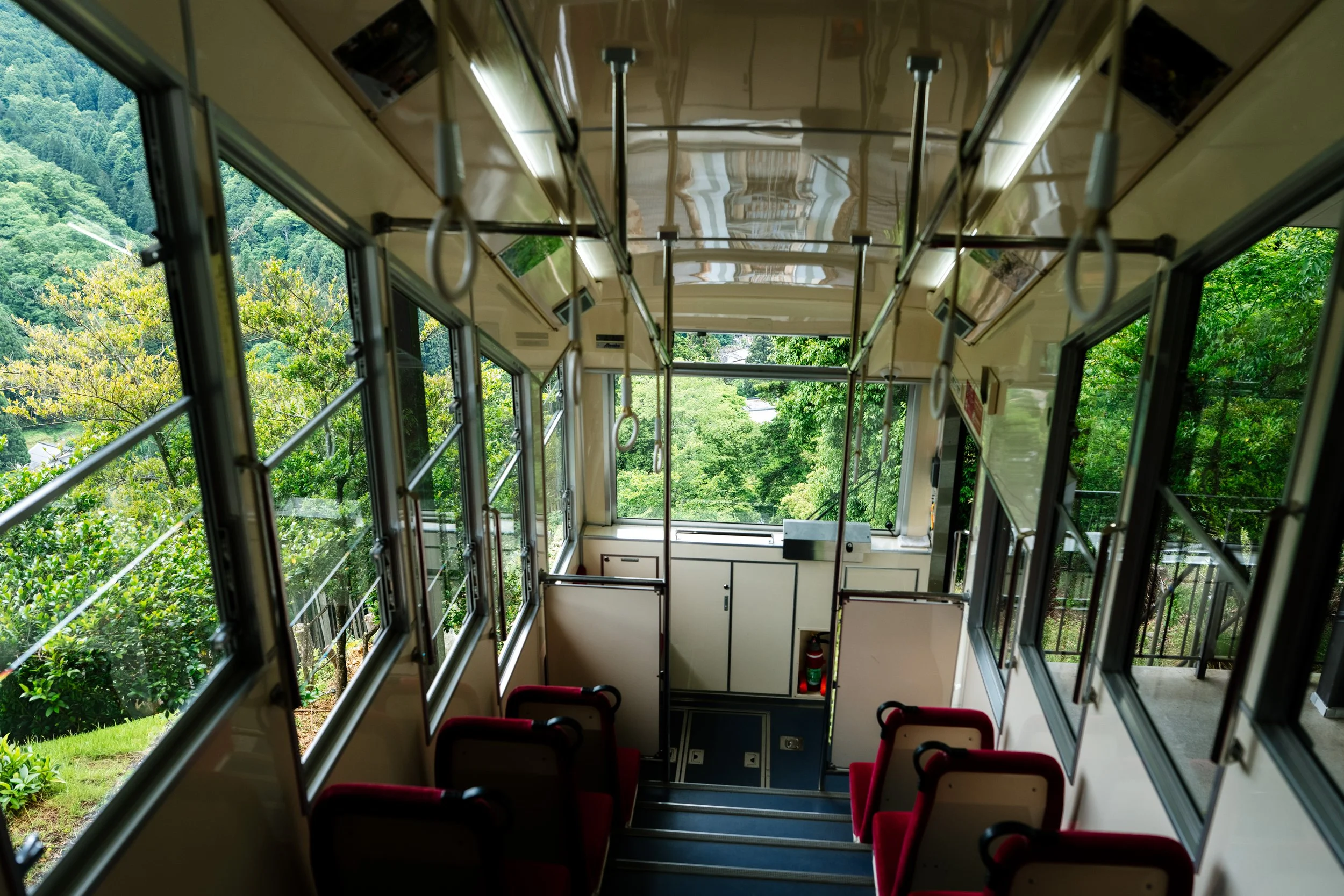 Interior view of a funicular or cable car showing red seats and large windows with a view of green trees and hillside outside.