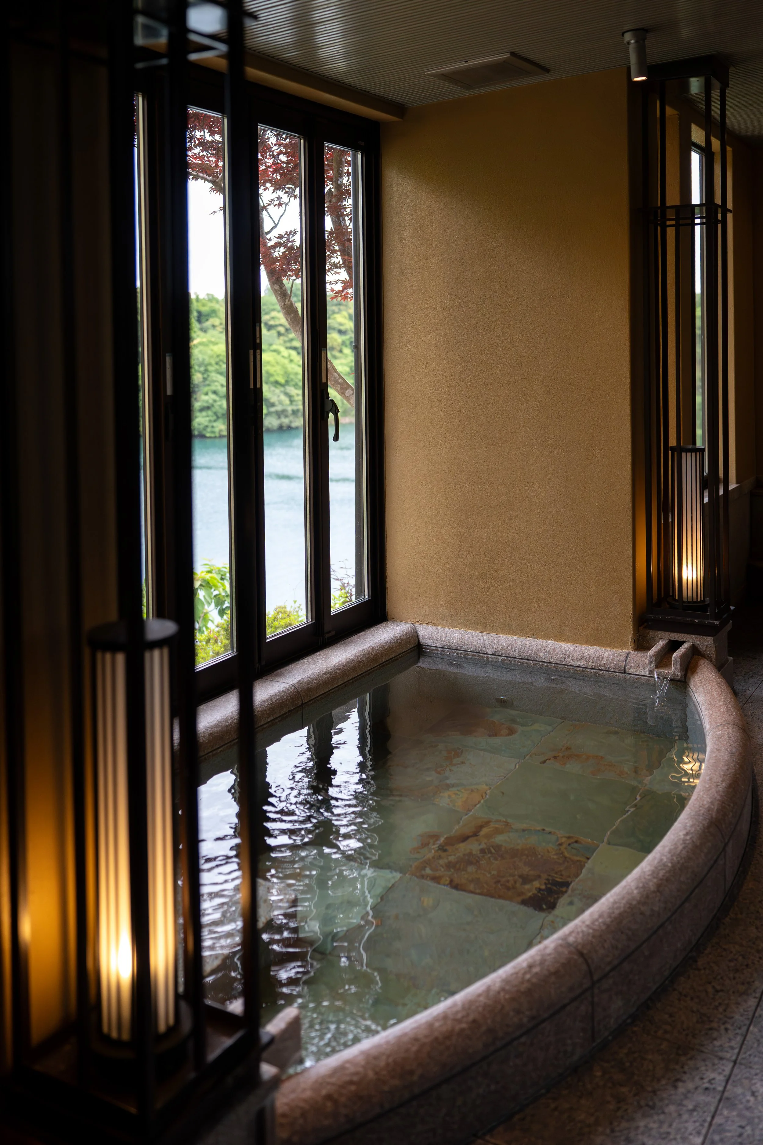 Indoor hot spring bath with large window showing trees and water outside, decorated with warm lighting and traditional Japanese design elements.