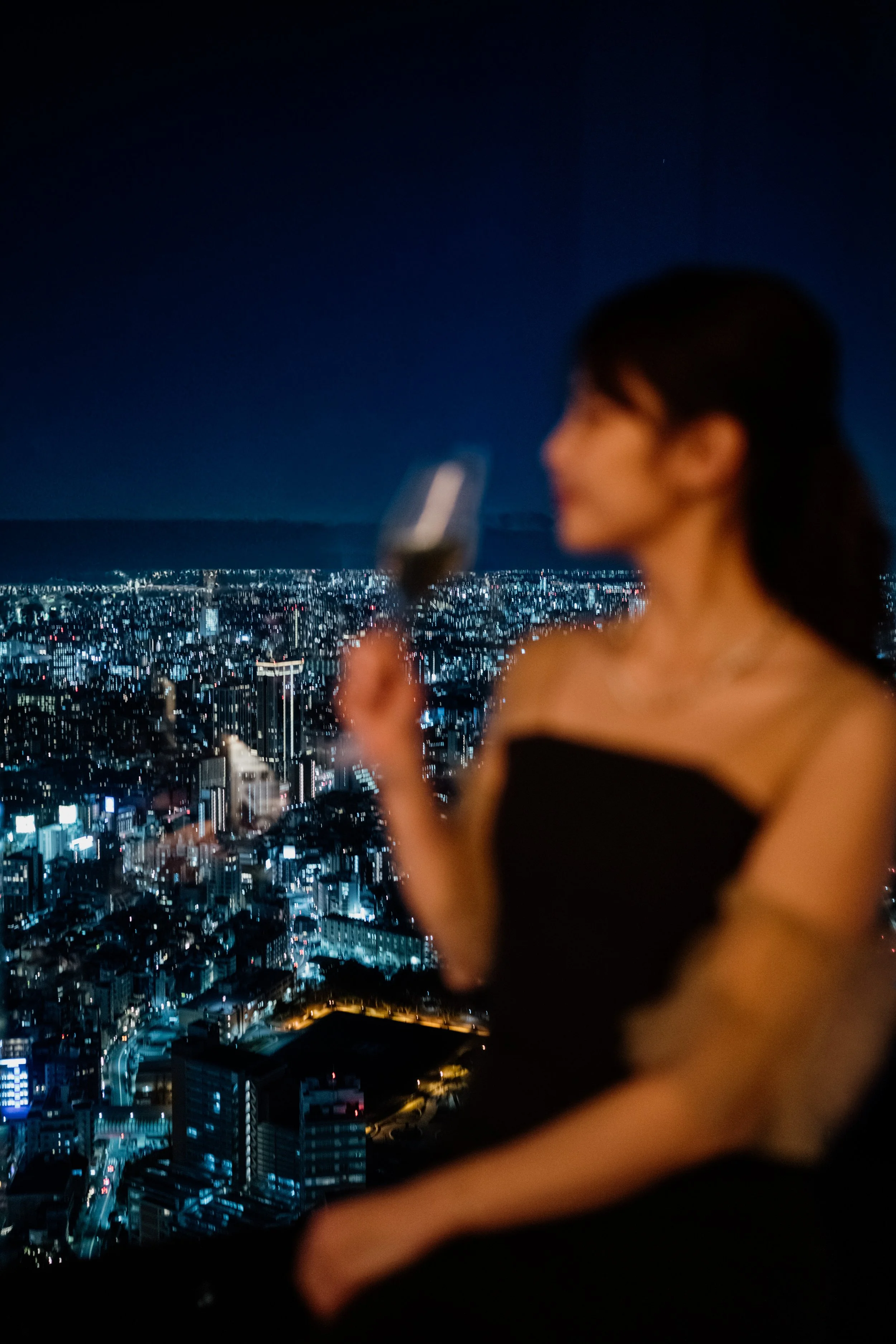 A woman in a black strapless dress is looking at her smartphone against a cityscape at night, with illuminated buildings and a dark sky at The Ritz-Carlton, Tokyo. Editorial hotel photography, Japan.