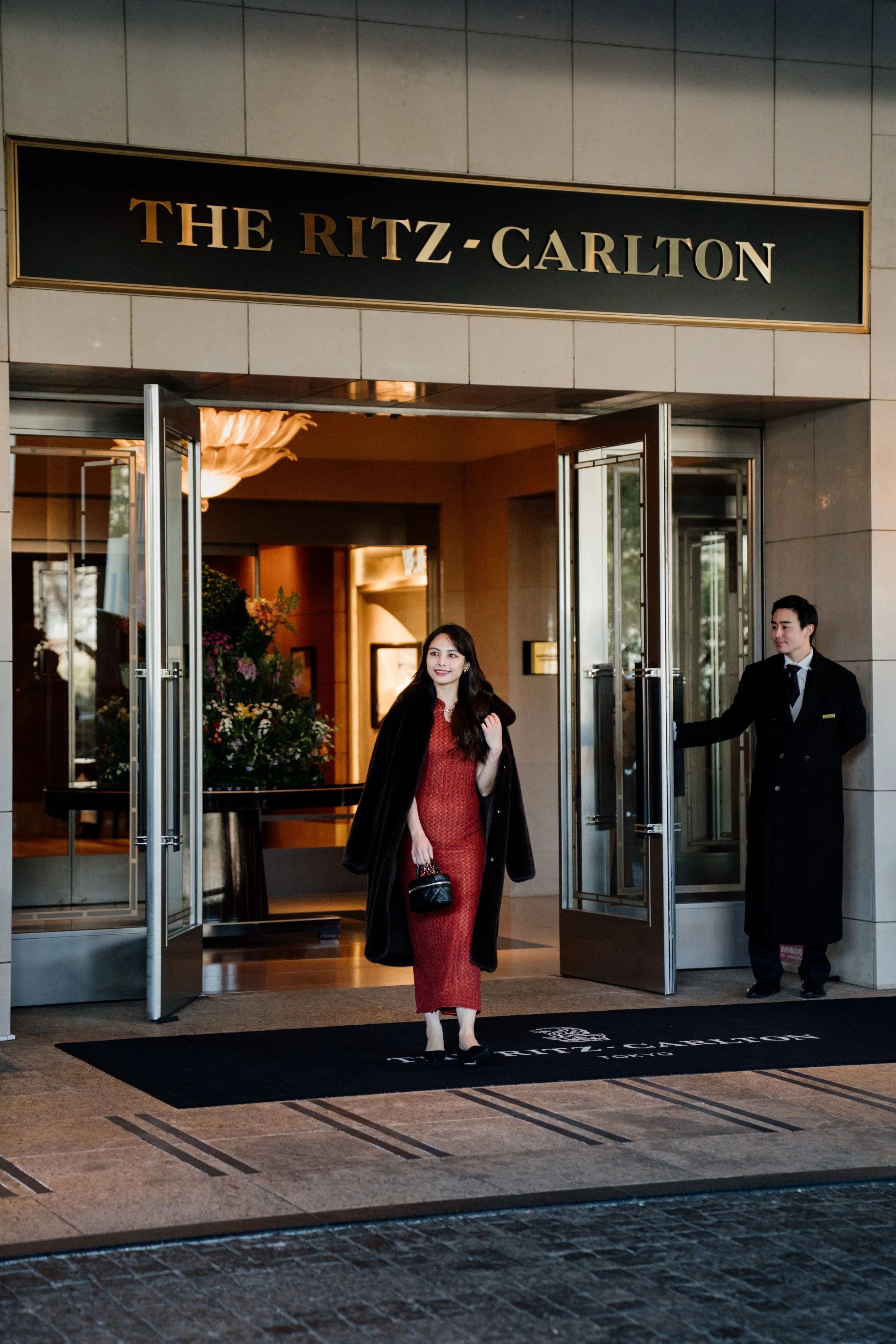 A woman with long dark hair wearing a red dress and black coat exiting The Ritz-Carlton hotel, with a doorman holding the door open behind her, inside the hotel lobby with floral arrangements and warm lighting. Editorial hotel photography, Japan.