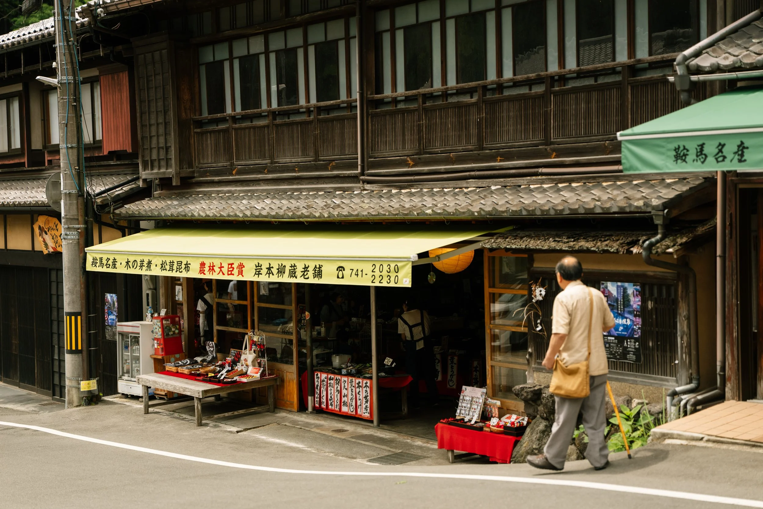 A street scene featuring a traditional Japanese building with a small shop outdoors offering various souvenirs and artifacts. Two people are walking past, one with a cane and the other wearing a yellow coat. The building has wooden railings and a til