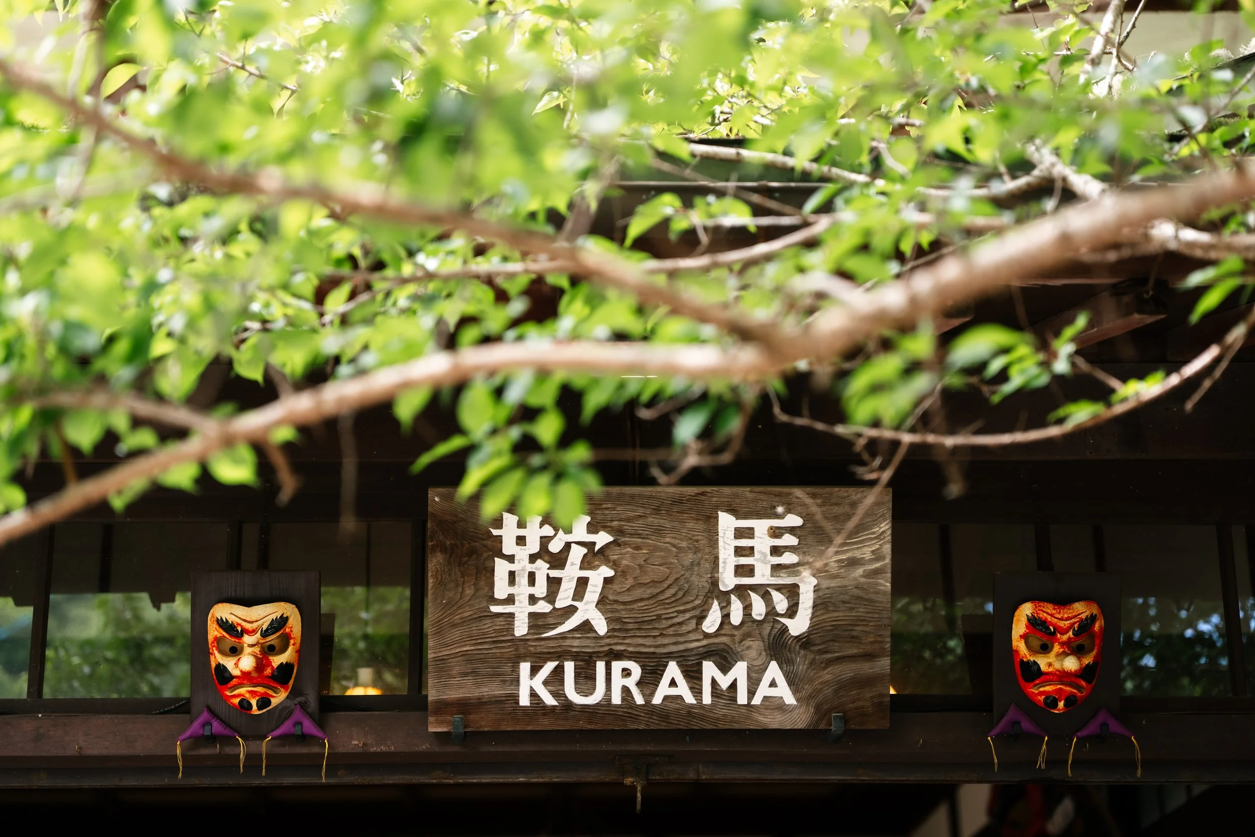 A wooden sign with white Japanese characters and the word "KURAMA" beneath it, hanging below green foliage, with two traditional Japanese masks flanking the sign.