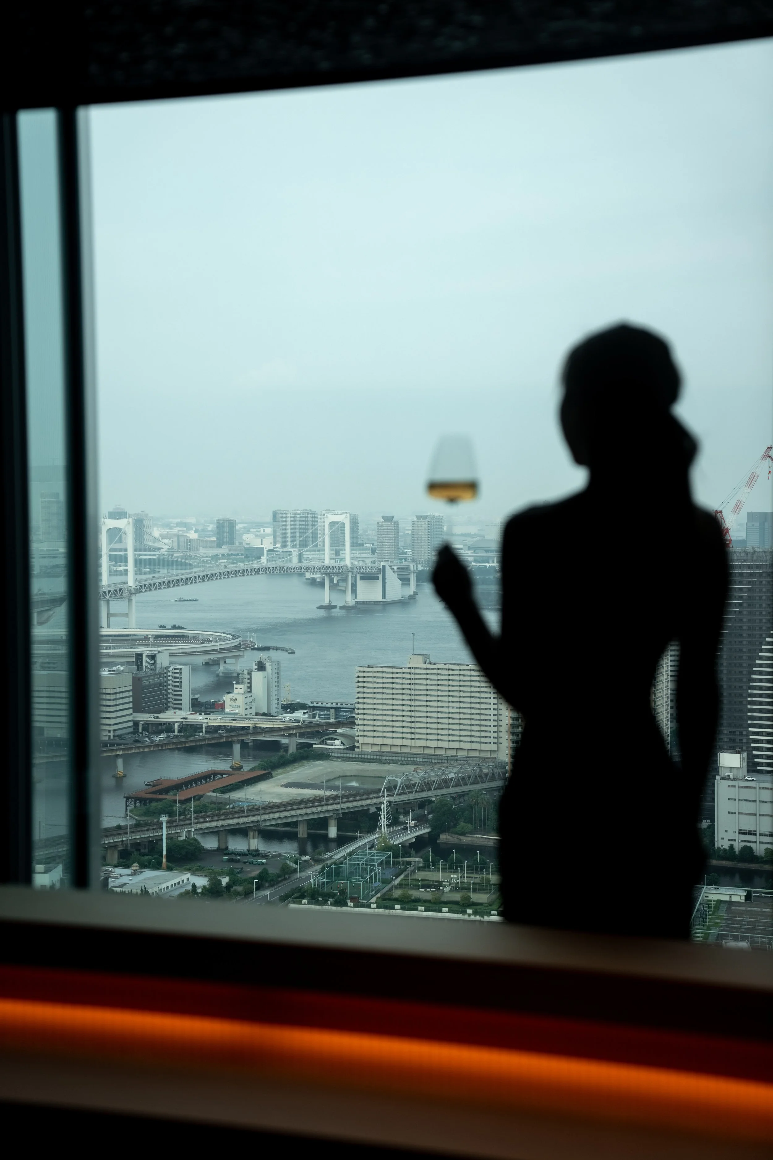 A silhouette of a person wearing glasses, looking out a large window at a cityscape with bridges over a river and tall buildings under a cloudy sky at JW Marriott Tokyo hotel, editorial hospitality photography, Japan.