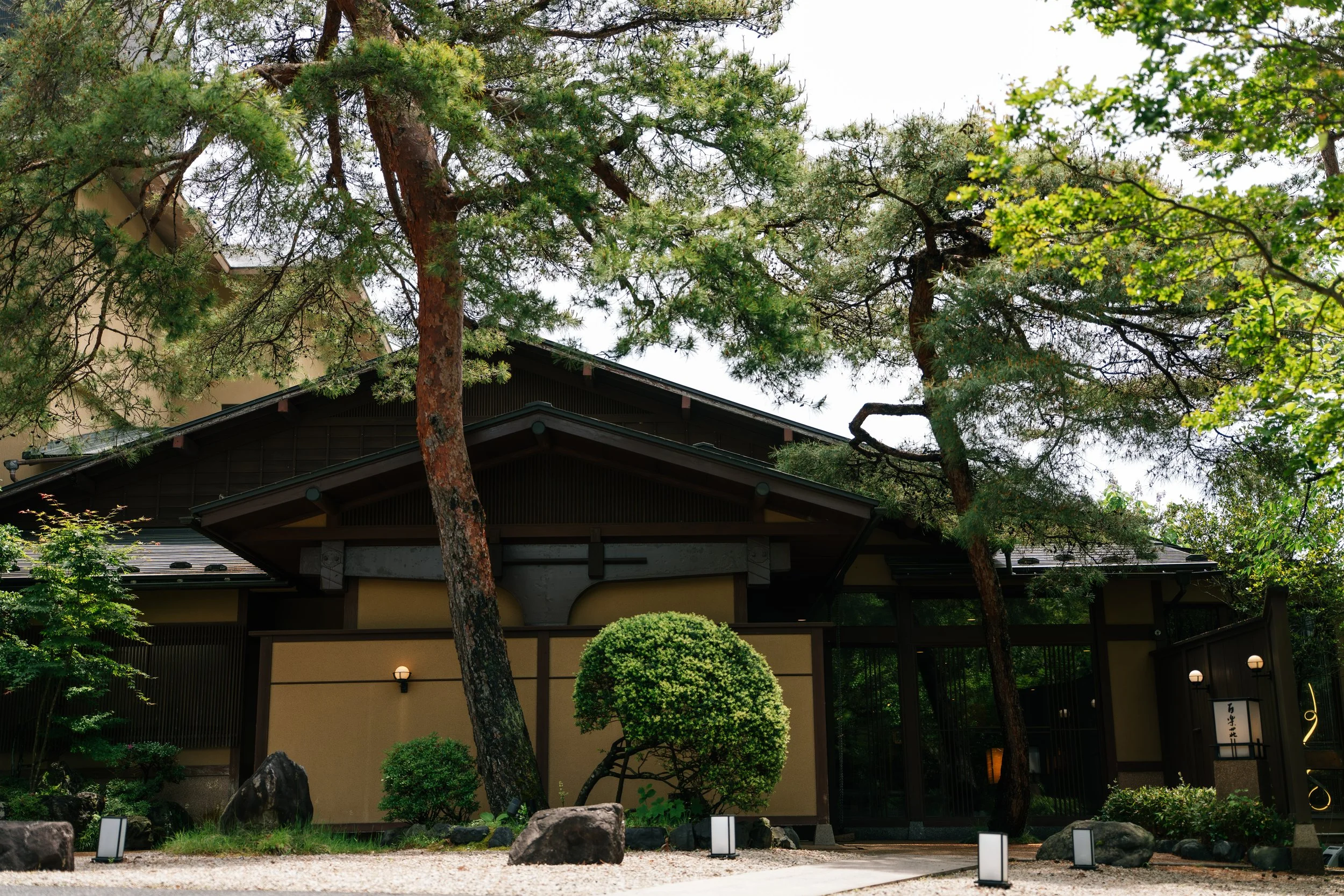 Traditional Japanese building surrounded by green trees and shrubs, with rocks and pathway in the foreground, illuminated lanterns, and a sign with Japanese characters.