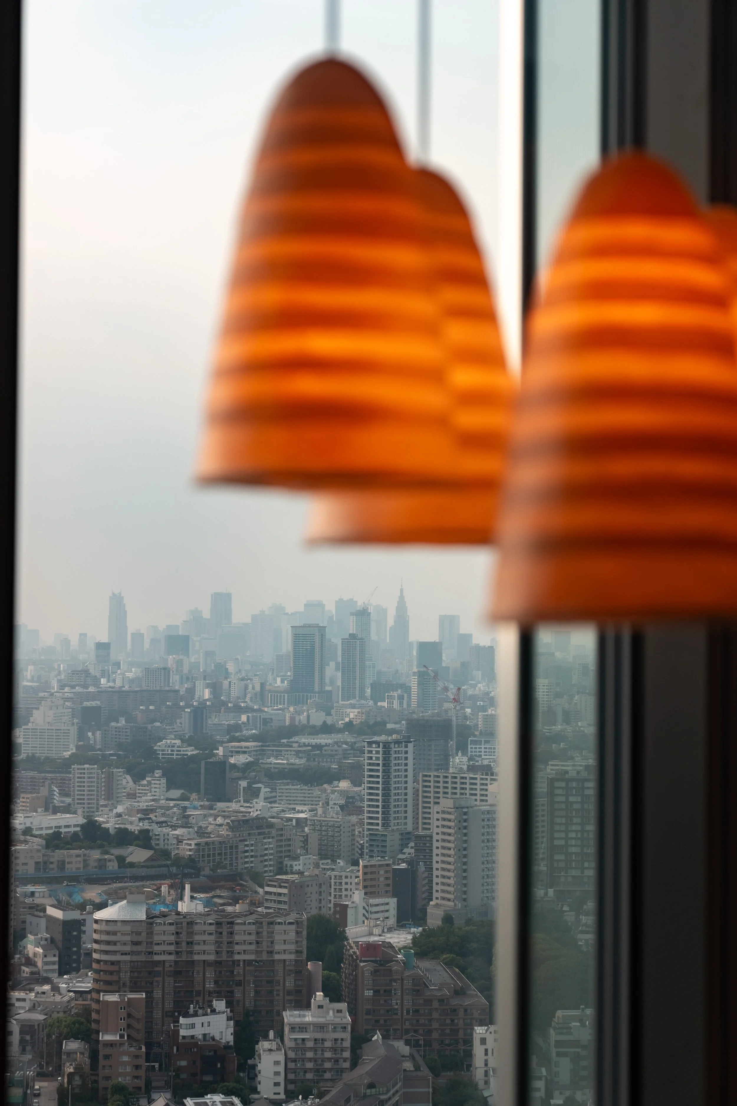 View of a city skyline with tall buildings and skyscrapers seen through a window, with orange-striped pendant lights hanging in the foreground at JW Marriott Tokyo hotel, editorial hospitality photography, Japan.