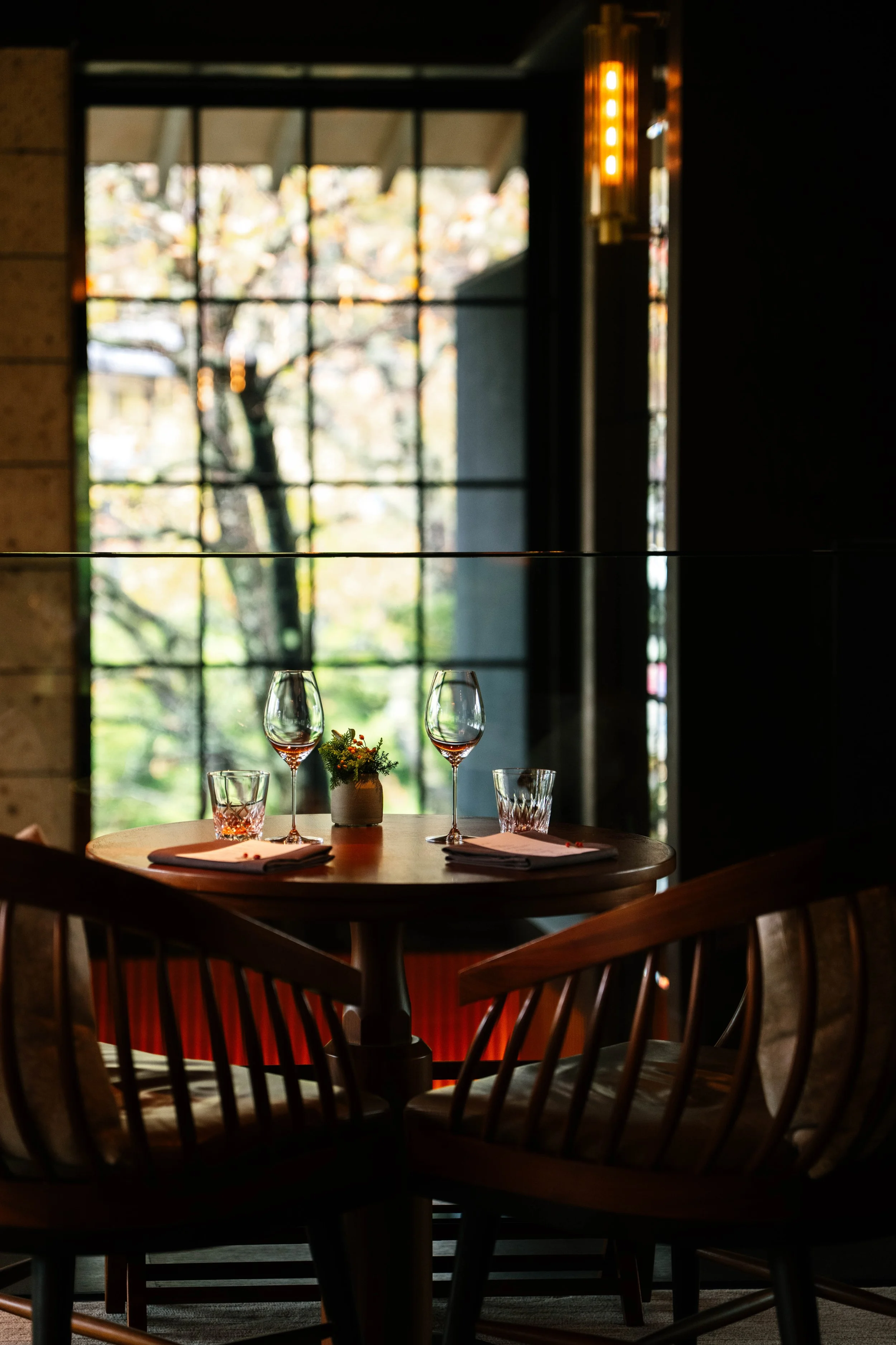 Table set for two with wine glasses, a small centerpiece, and napkins in a cozy restaurant with large window showing trees outside.