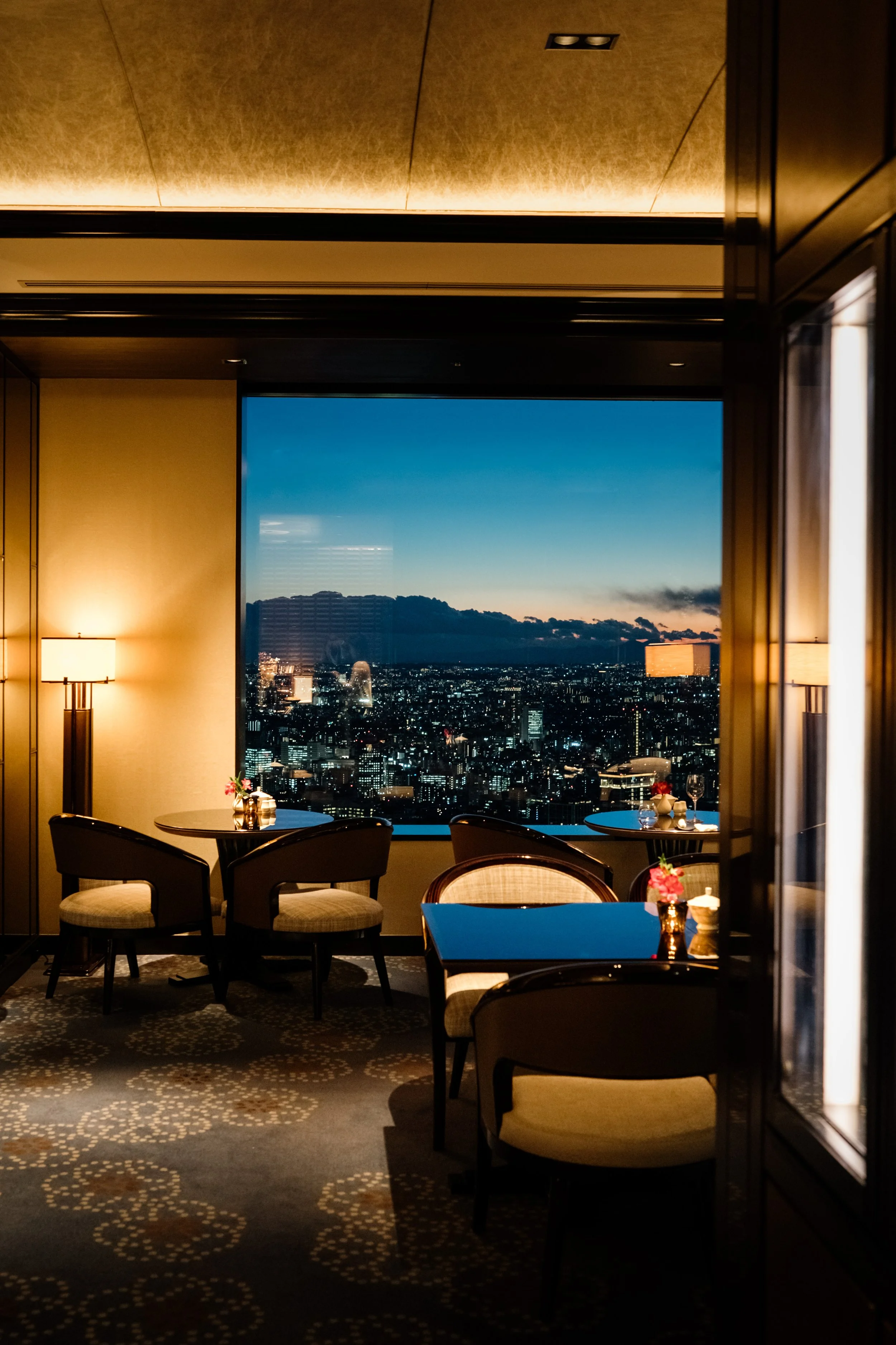 Interior of a high-rise restaurant or lounge with a large window showing a city skyline at dusk, tables set with small vases of pink flowers, and chairs with upholstered seats.