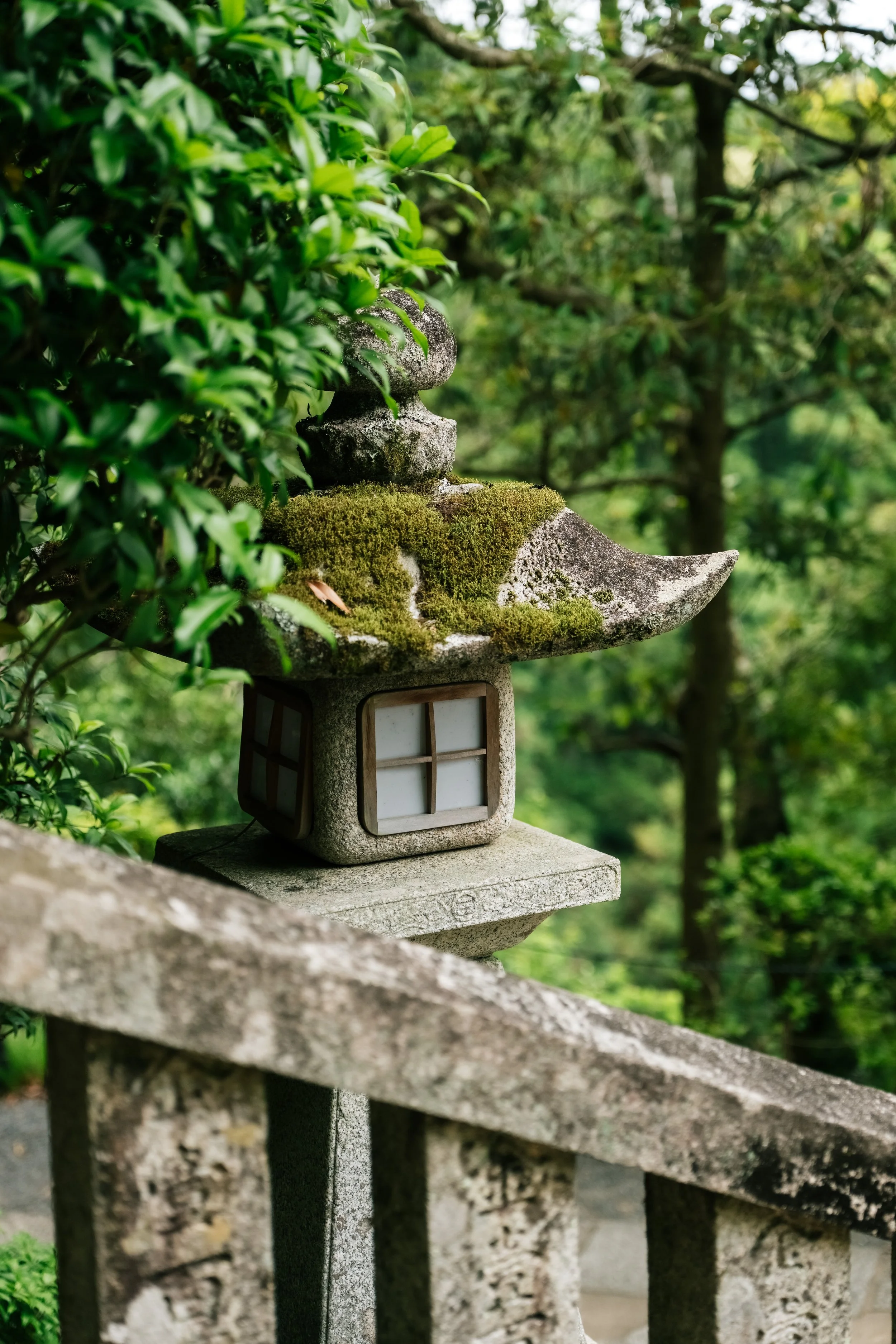 Stone lantern with moss on top, located beside a stone railing in a lush green garden, surrounded by trees and foliage.