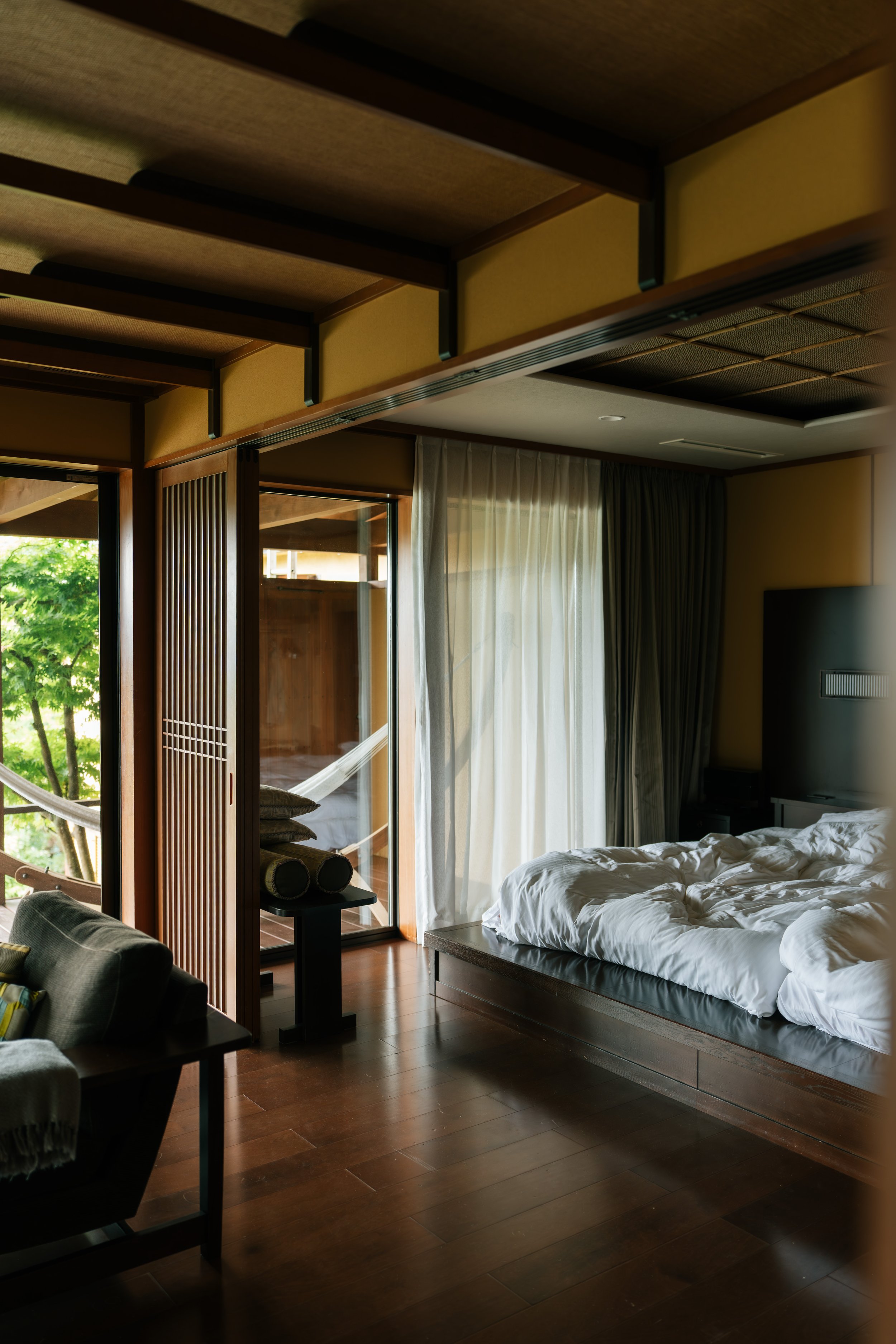 A bedroom with a sliding glass door leading to an outdoor balcony, a bed with white bed linens, and a window with sheer and dark curtains. There is wooden ceiling detail and a hammock visible outside.