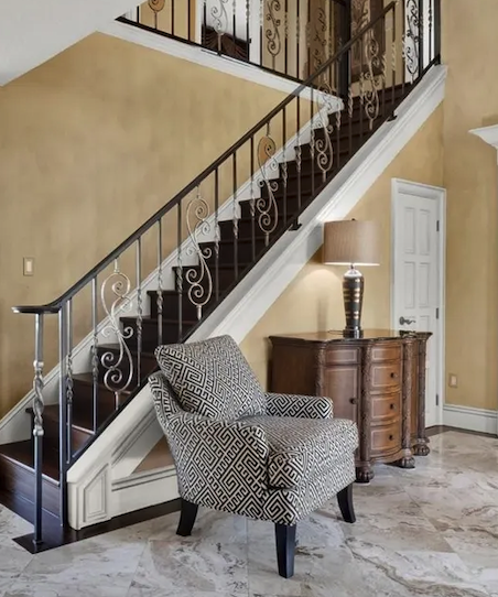 A cozy corner with an armchair, a side table with a lamp, and a staircase with decorative wrought iron railing in a house interior.