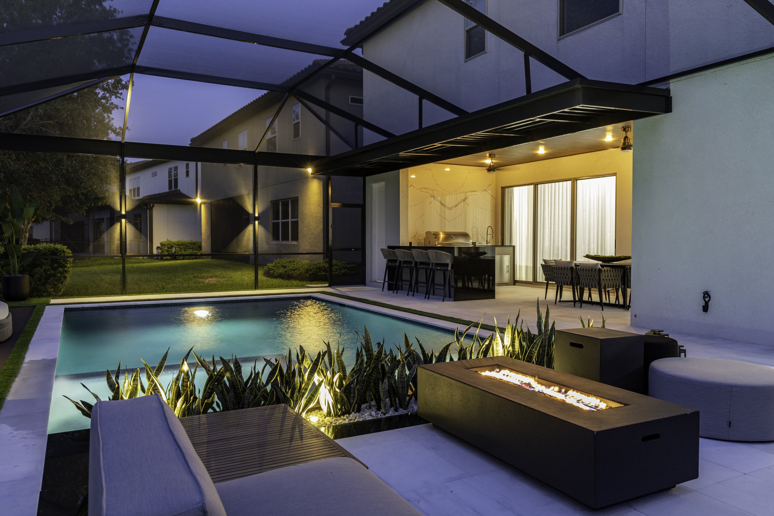 Modern outdoor living design with swimming pool, fire feature, custom seating and outdoor kitchen illuminated at night.