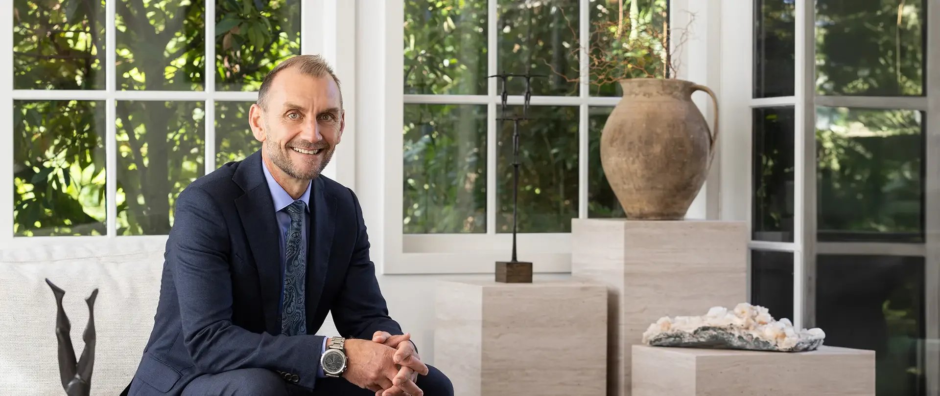 A smiling man in a dark suit and watch sitting indoors near large windows with greenery outside, with decorative vases and sculptures in the background.