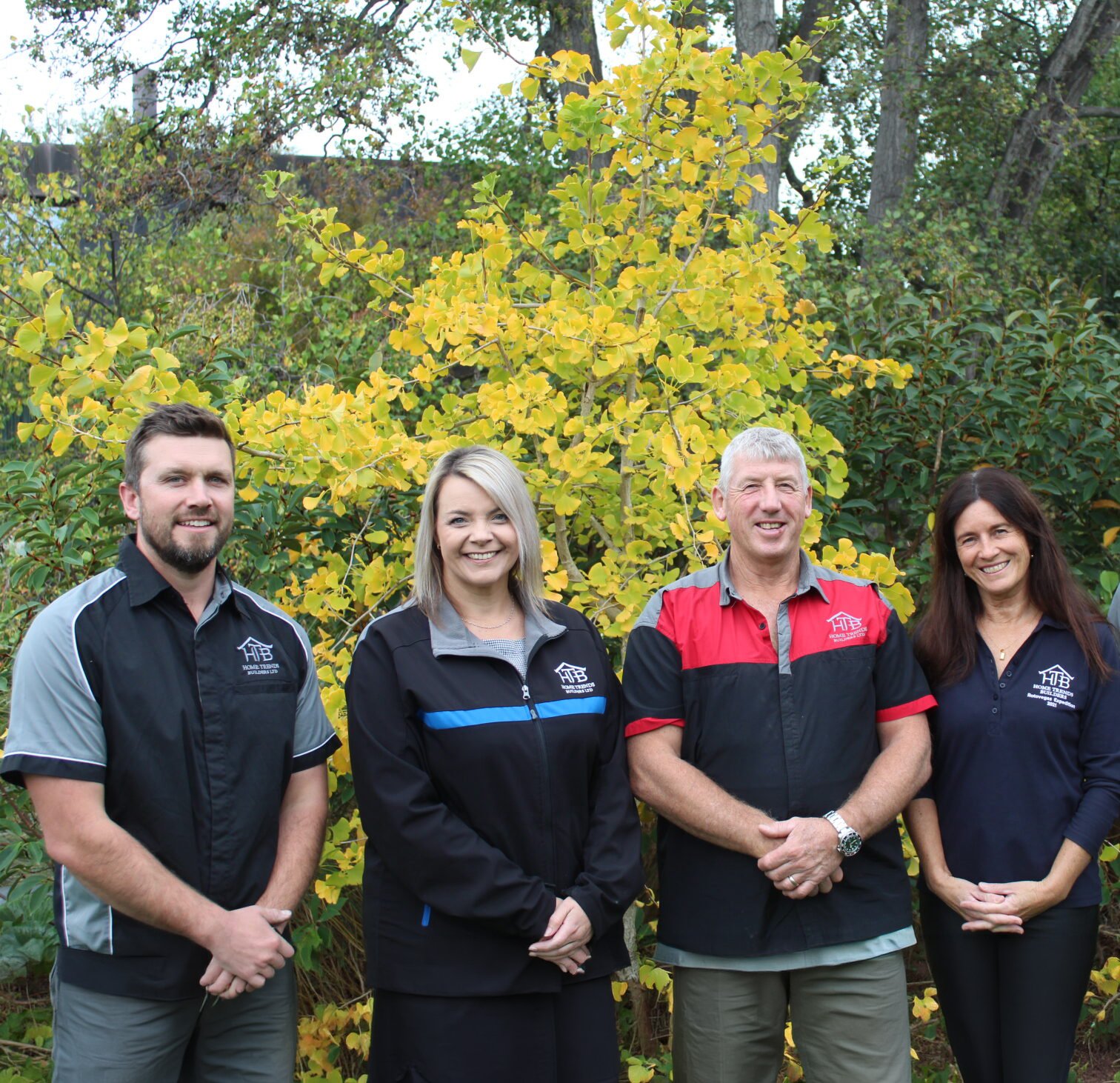 Group of four people standing outdoors in front of a yellow-leaved tree, smiling and wearing black, blue, red, and navy team shirts with a logo.