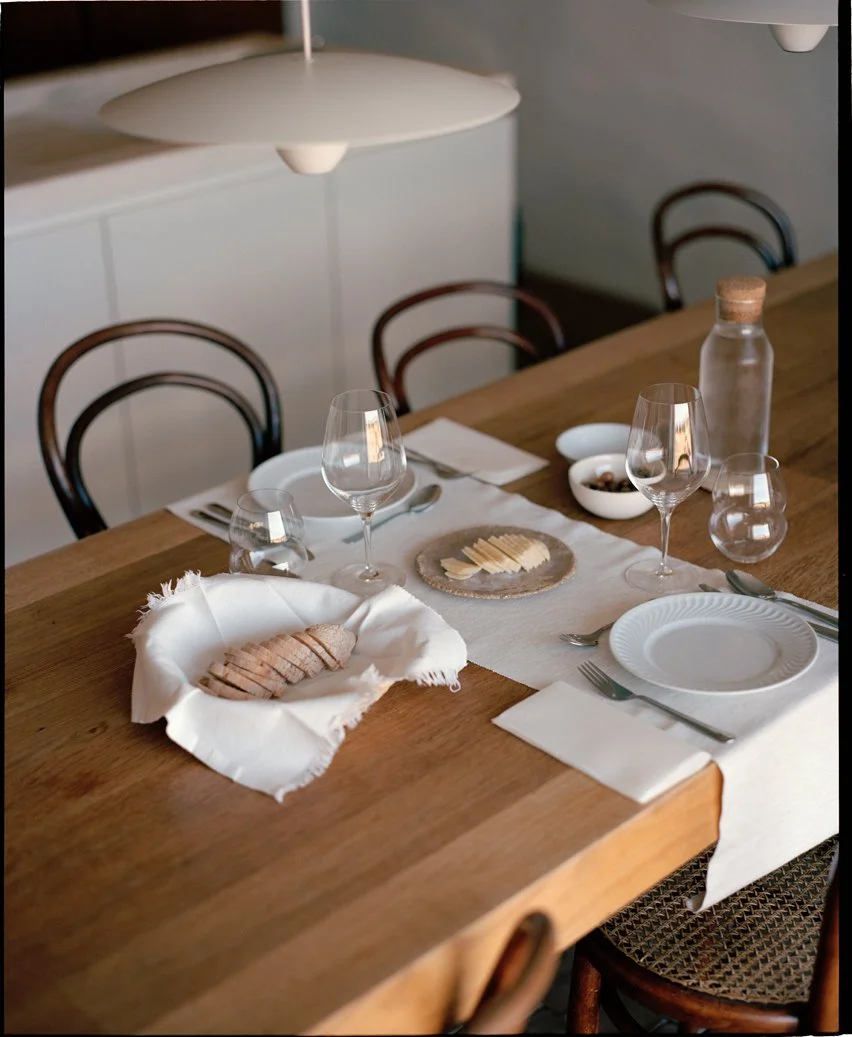 A neatly set dining table with white plates, wine glasses, silverware, and a cloth napkin with sliced bread. There are small bowls with condiments or spreads and a glass bottle with a cork. The table has a white table runner, and the background shows chairs and a hanging light fixture.
