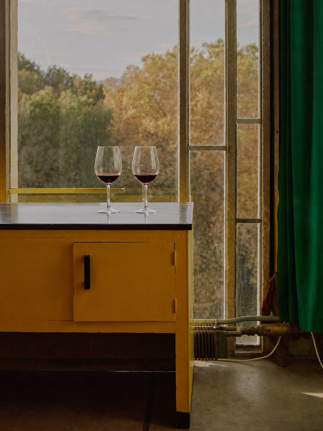 Two glasses of red wine on a yellow table near a large window with green curtains, overlooking autumnal trees.