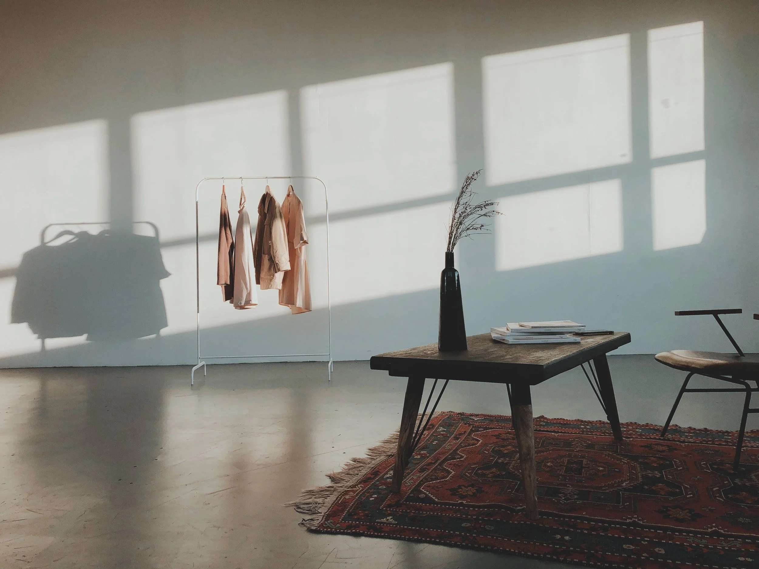 A minimalist interior with sunlight casting rectangular shadows on a white wall, a white clothing rack with neutral-colored clothes, a tall black vase with dried branches, a wooden table with a stack of magazines, and a patterned rug.