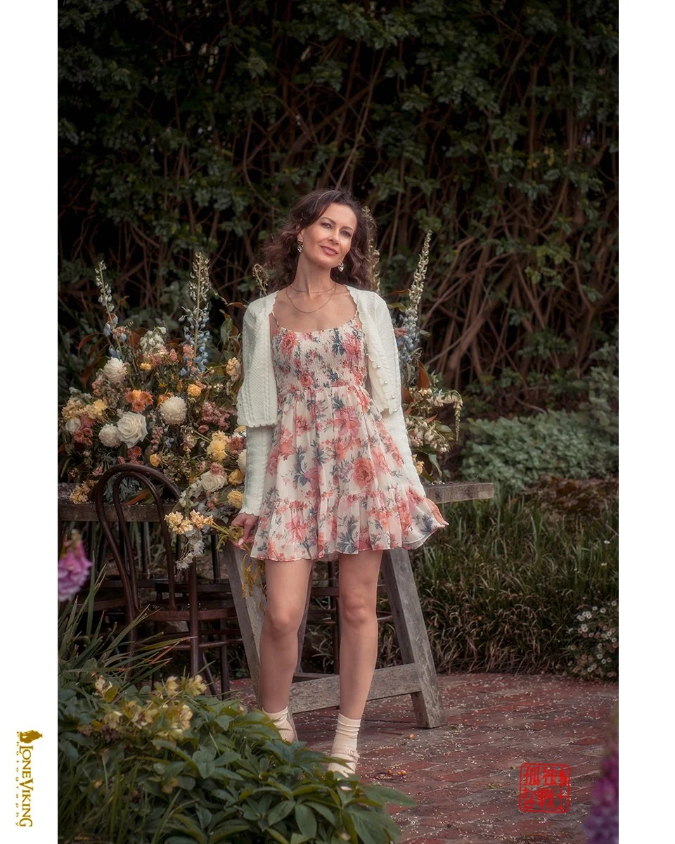 A woman standing outdoors in front of green leafy bushes, wearing a floral dress, off-white cardigan, and cream socks with shoes. She is beside a garden bench and surrounded by various flowers.